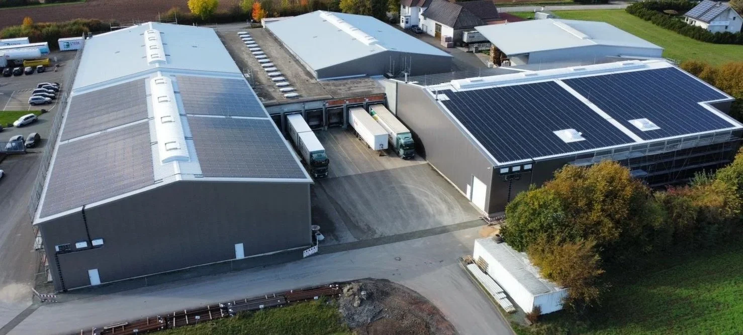 Luftaufnahme eines Industriegebäudes mit Solaranlagen auf den Dächern, mehreren Parkplätzen und Lkw-Ladezonen.