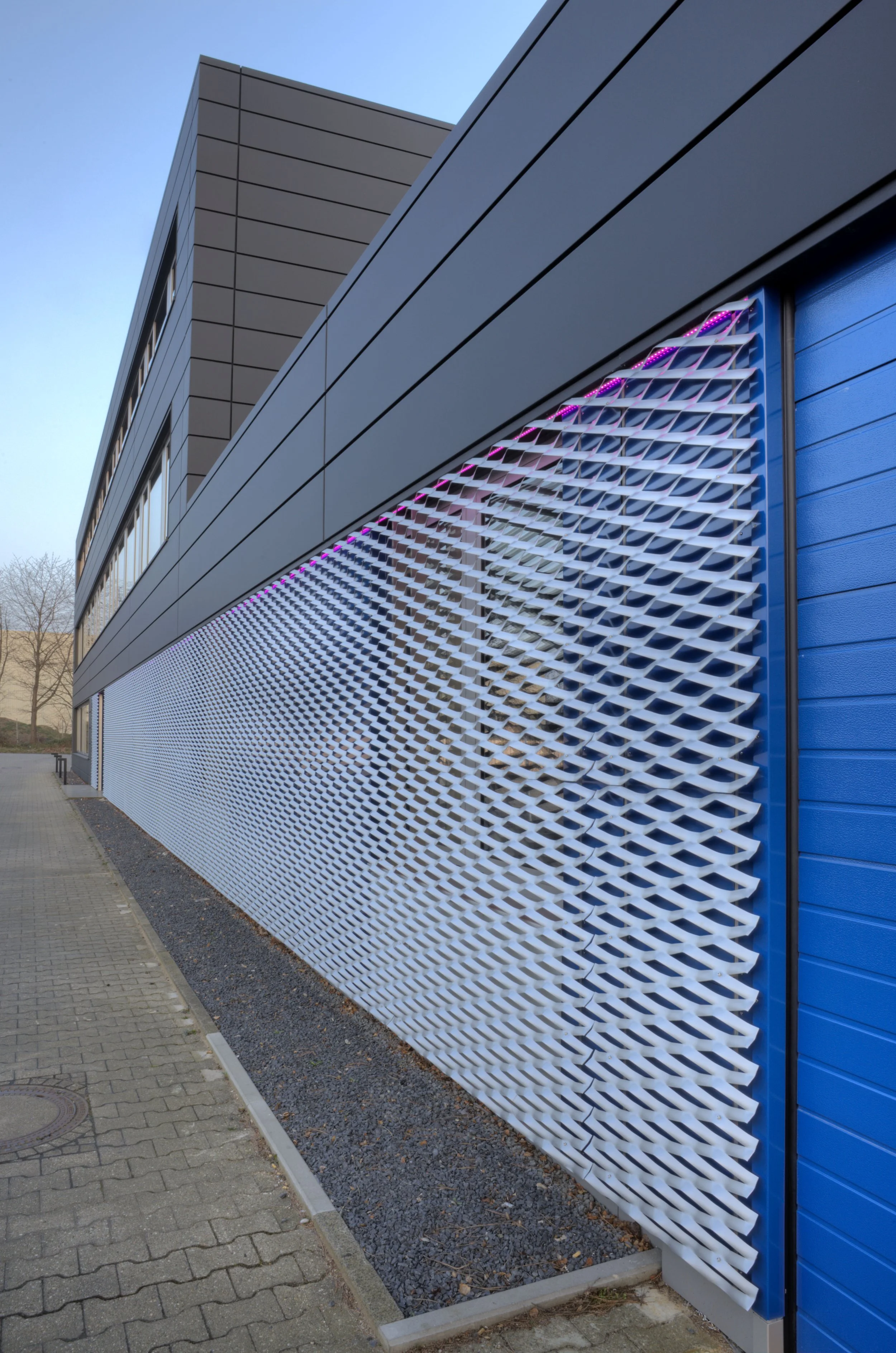 Modernes Gebäude mit einer sichtbaren blauen Wand und einem innovativen weißen Gitter-Design an der Außenseite. Der Himmel ist klar und blau, und es gibt einen Gehweg im Vordergrund.