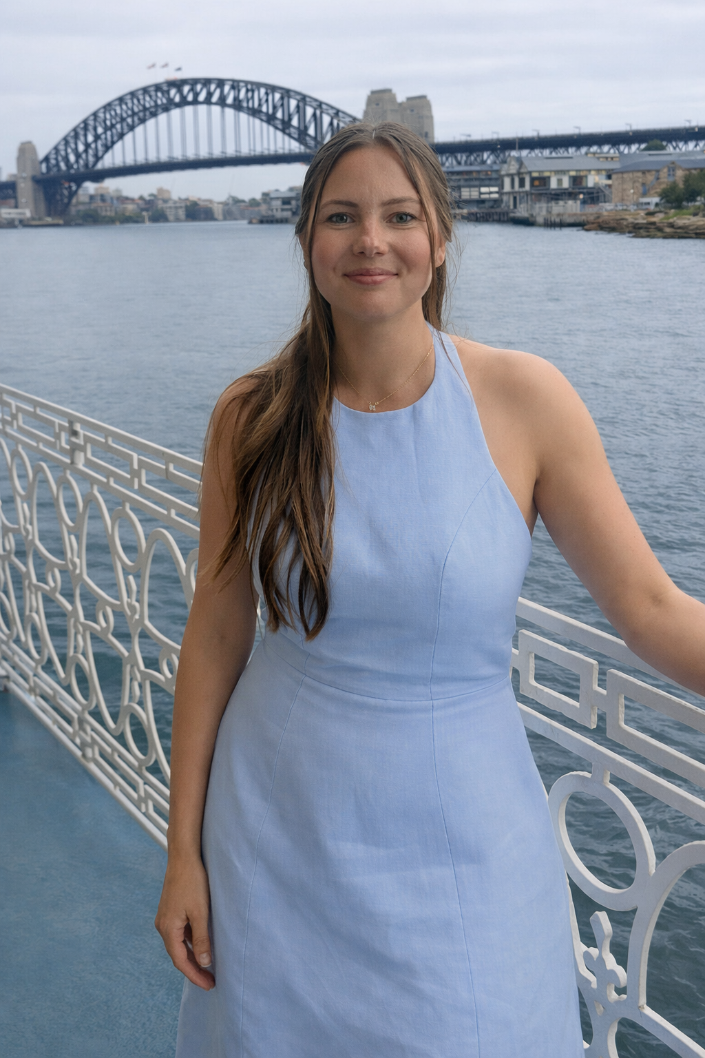 A woman in a light blue dress standing on a boat deck near a white railing, with the Sydney Harbour Bridge and city skyline in the background on a cloudy day.