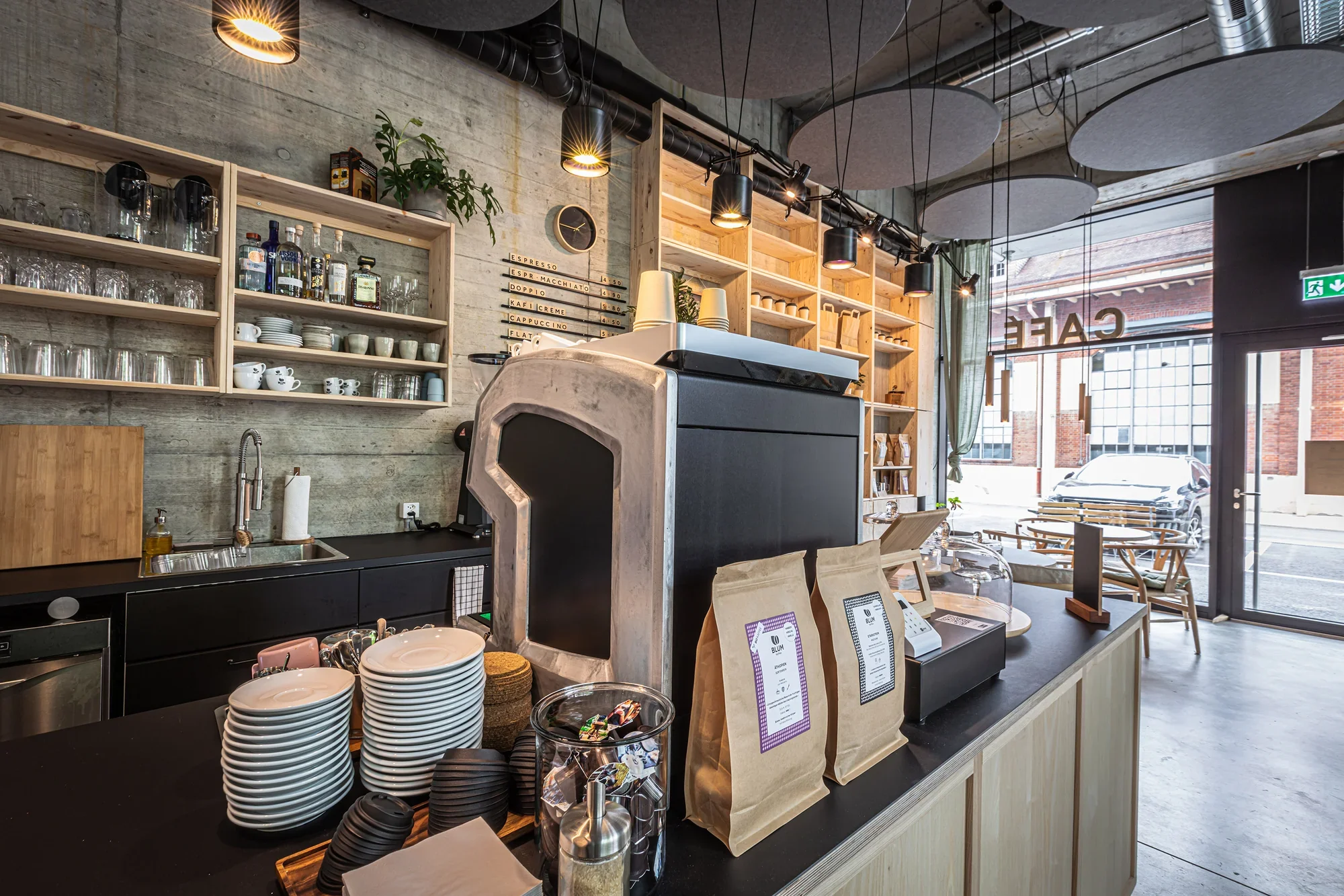 Inside a modern coffee shop with wooden shelves, a coffee counter, and large front windows showing parked cars outside.