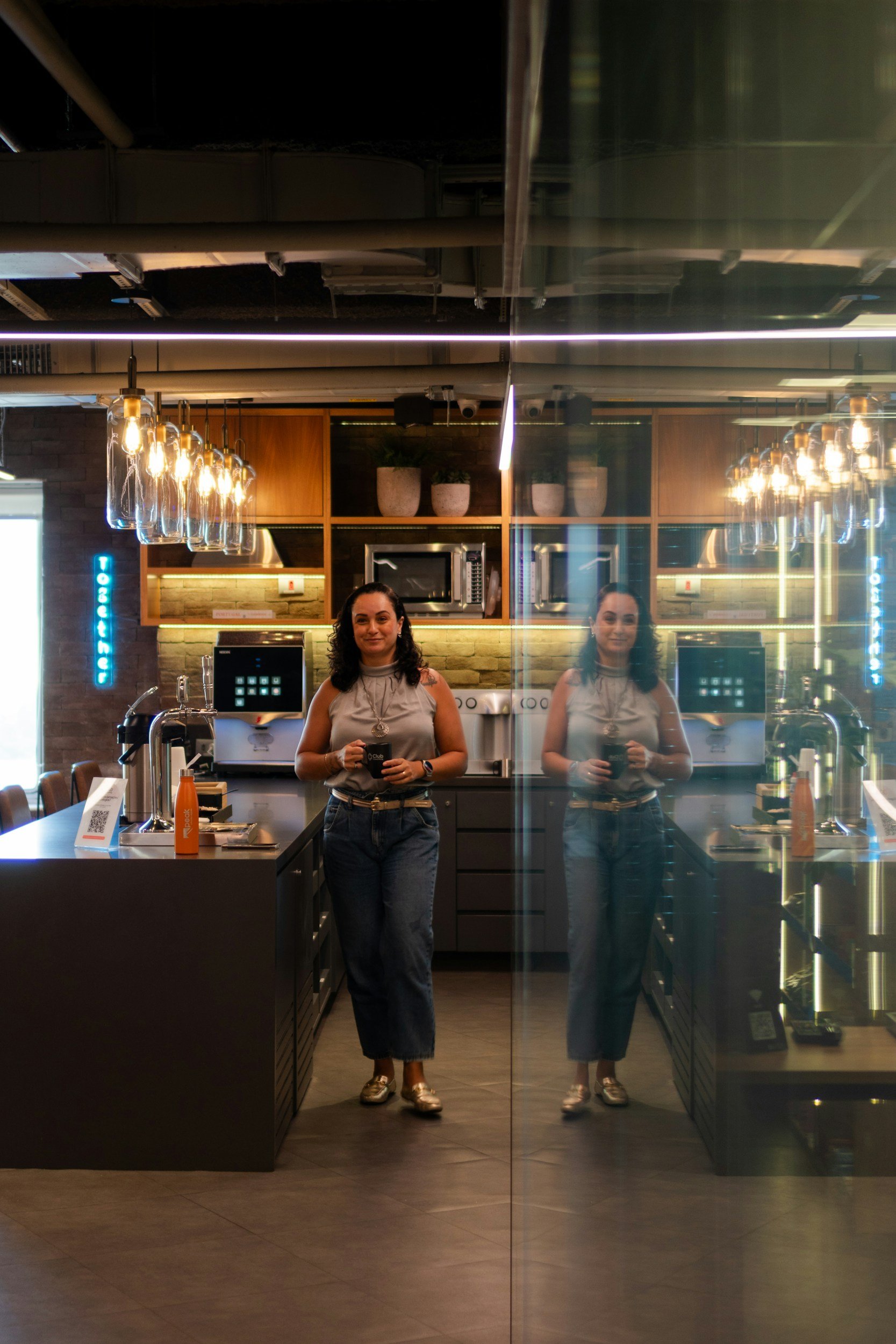 A woman standing behind a glass door in a modern coffee shop, holding a black mug, with her reflection visible on the glass, and a counter with coffee-making equipment and a visible menu.