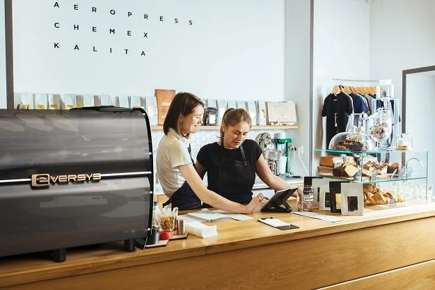 Two women working behind the counter at a coffee and baked goods shop, with a coffee machine and display case of pastries.