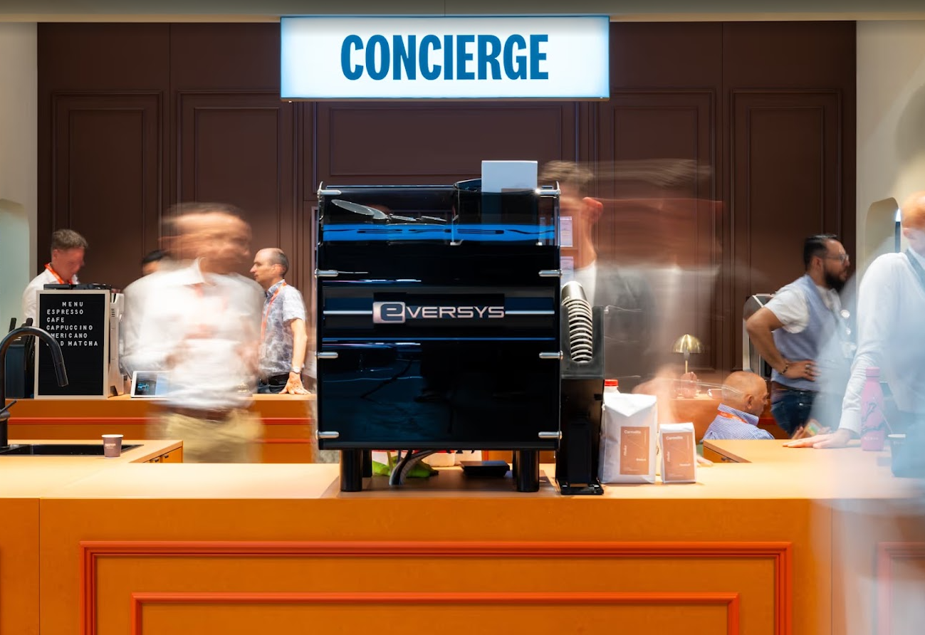 Coffee counter with a sign reading 'Concierge' above it, busy with blurred motion of people in line, a black Eversys coffee machine, and menu items for espresso, café, cappuccino, Americano, and matcha.