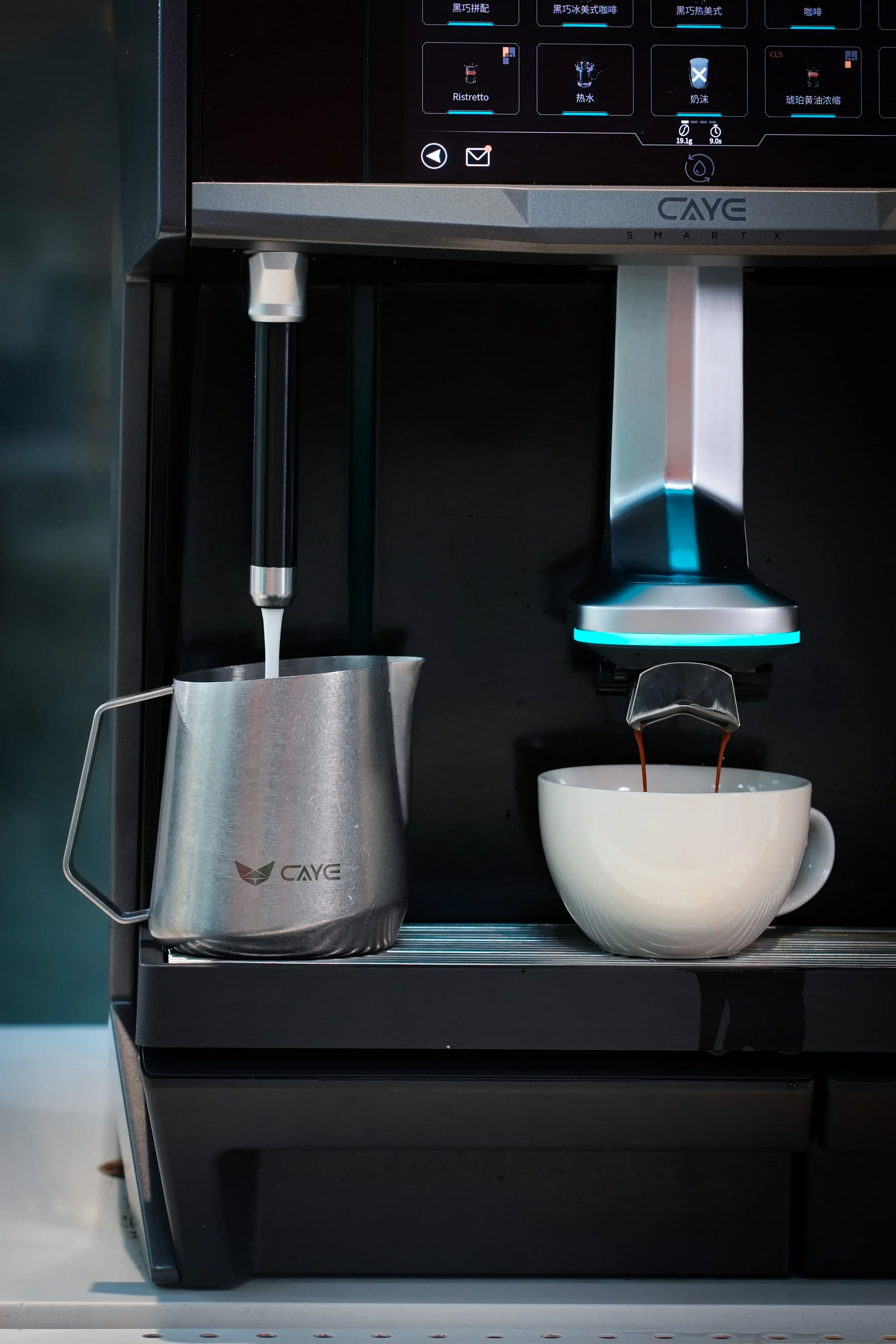 A CAYE coffee machine brewing coffee into a white mug, with a stainless steel milk pitcher positioned to the left of the mug.