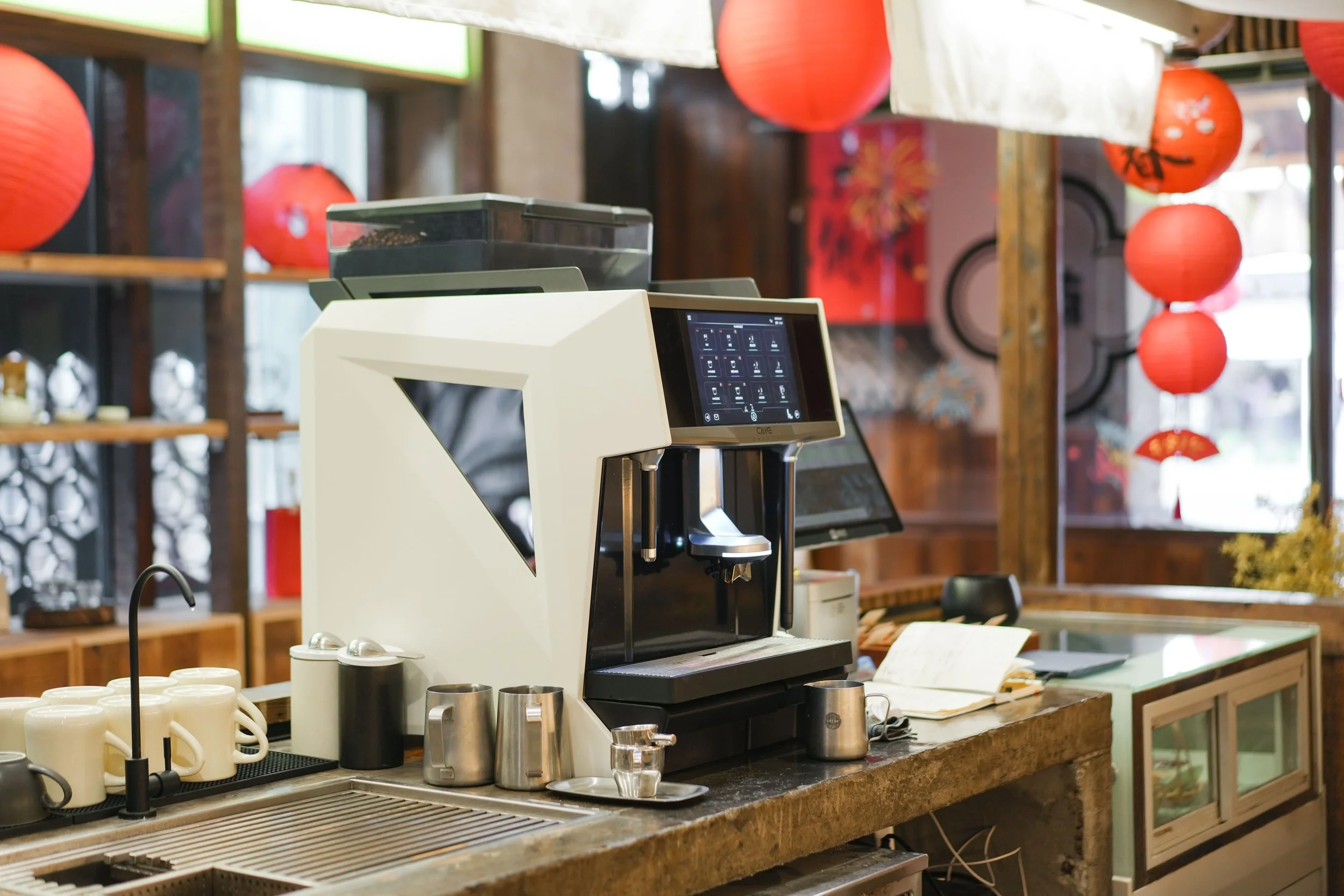 Automated CATE Coffee Rentals espresso machine on a counter with cups and ingredients, in a cafe with red paper lanterns and wooden shelves in the background.