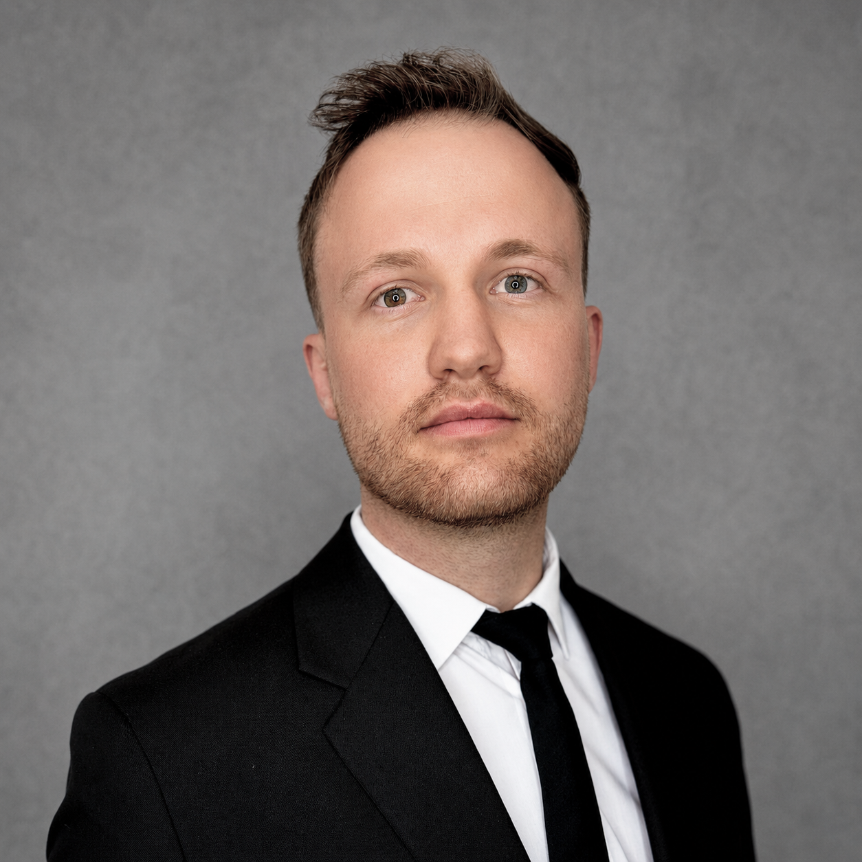 Headshot of a man with short brown hair, blue eyes, and light stubble, wearing a black suit and tie against a plain gray background.