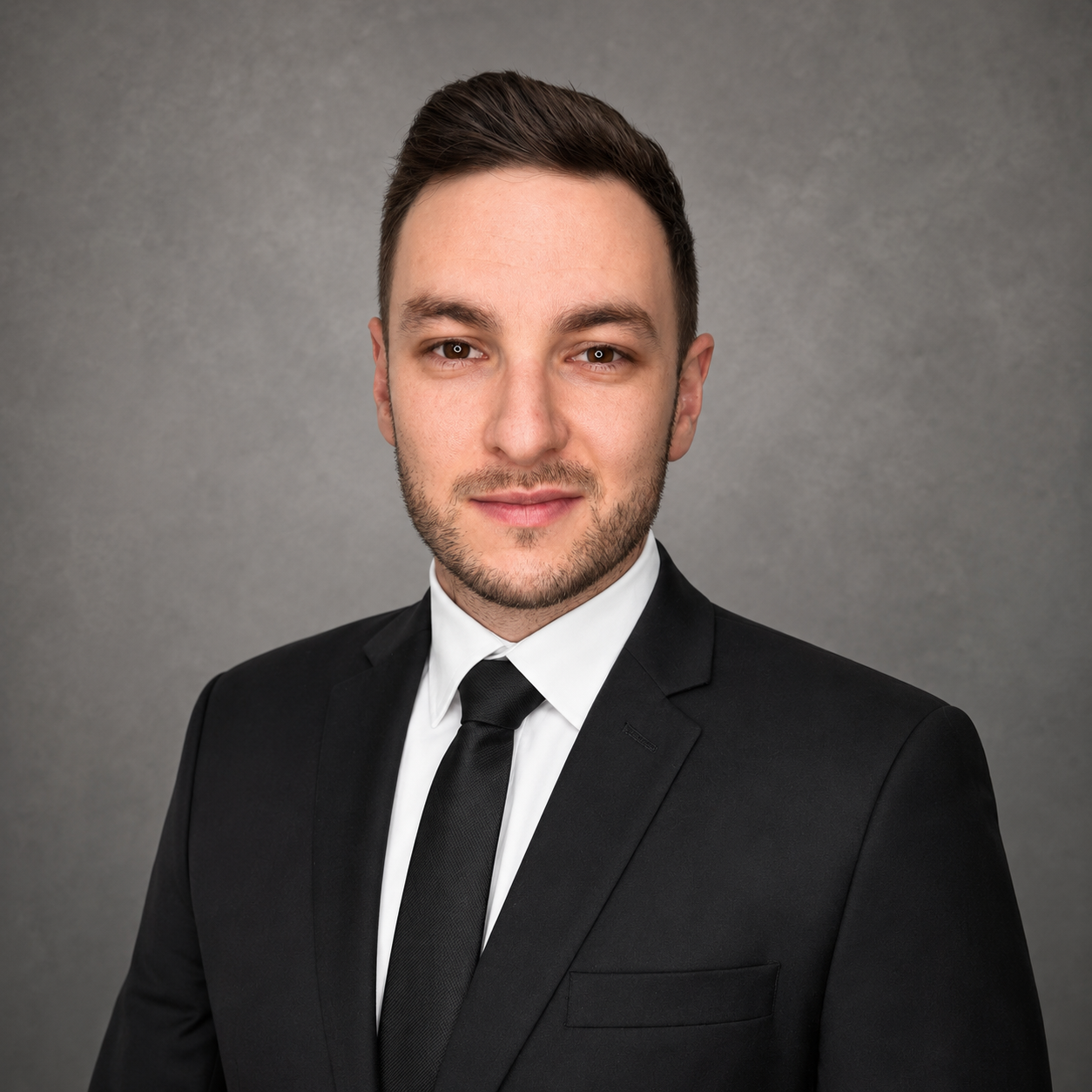 Young man in black suit, white shirt, and black tie posing against gray background.