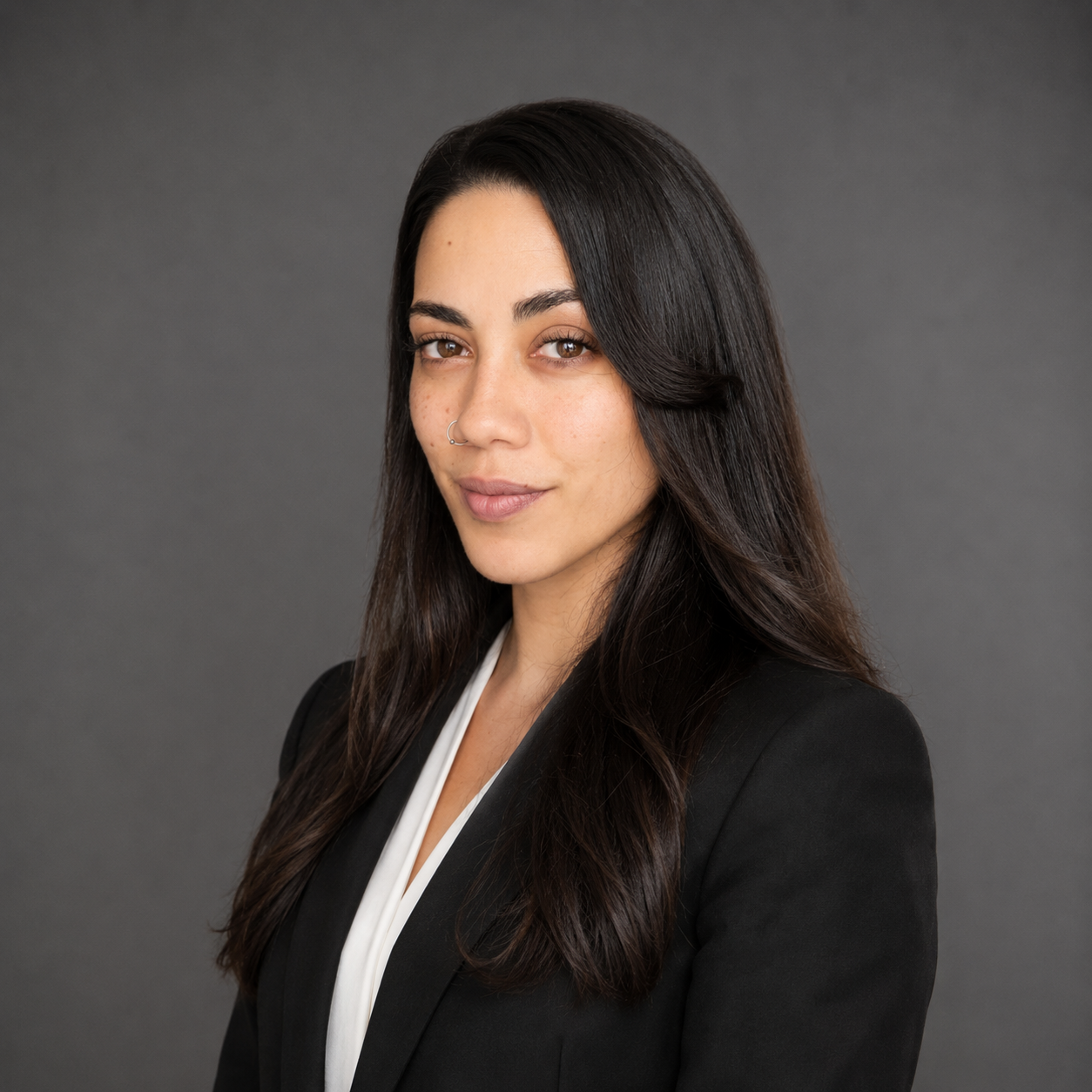 A woman with long dark hair, wearing a black blazer and white top, looking confidently at the camera against a gray background.