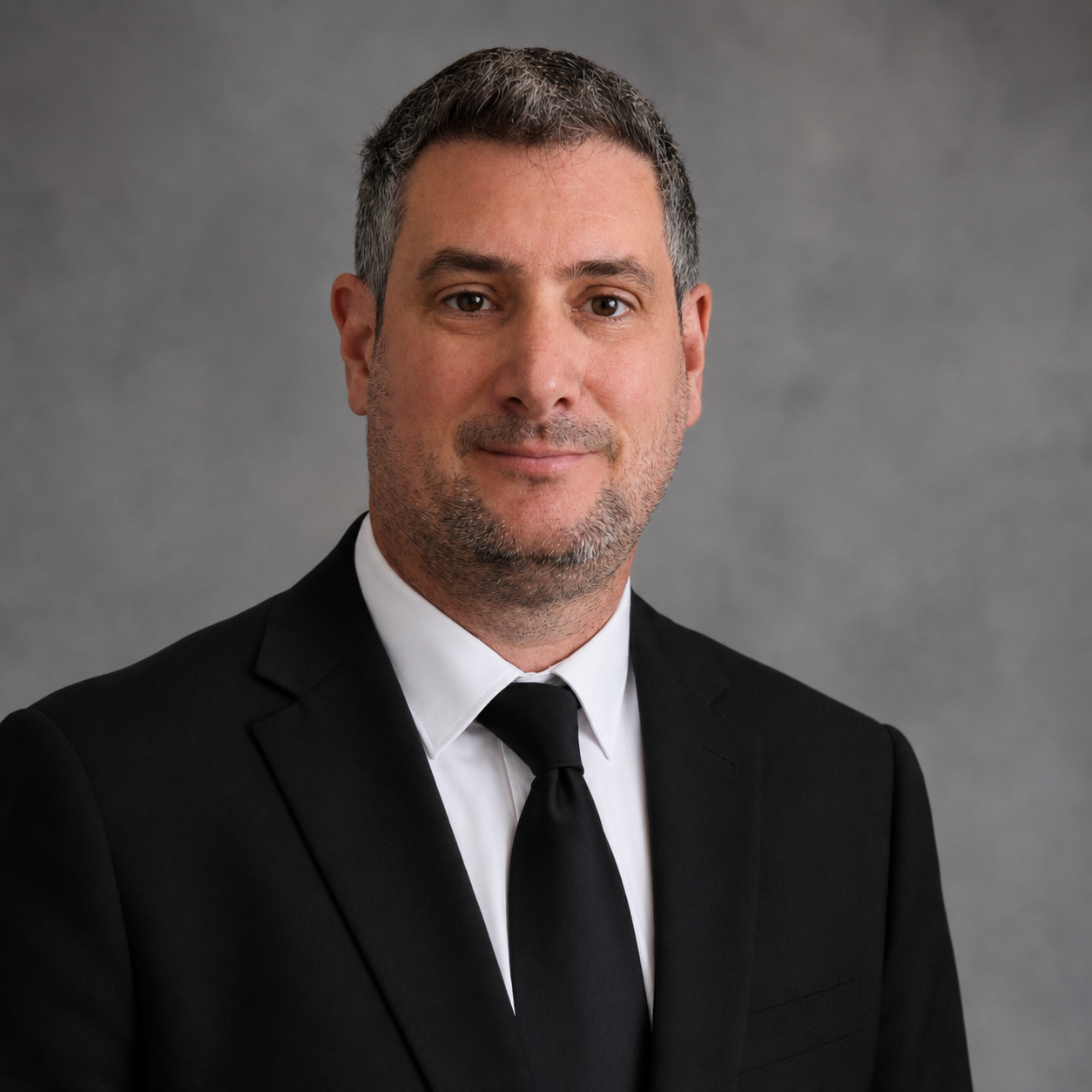 A professional portrait of a middle-aged man with short, salt-and-pepper hair, wearing a black suit, white shirt, and black tie, smiling with a neutral gray background.
