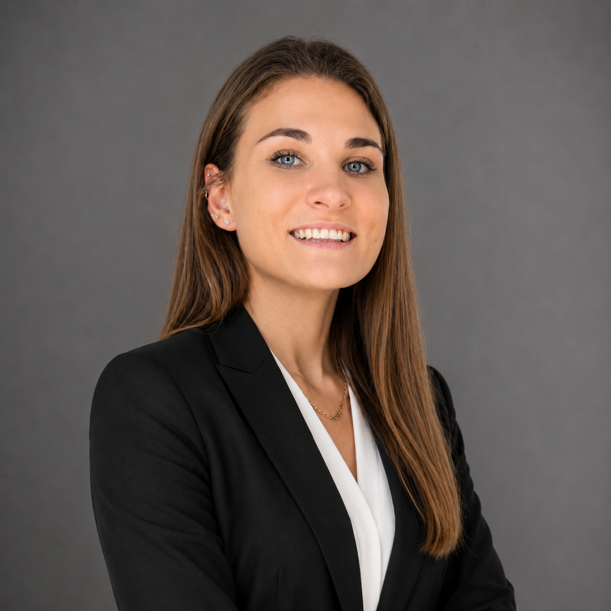 A young woman with long brown hair, blue eyes, and earrings, dressed in a black blazer and white blouse, smiling against a gray background.