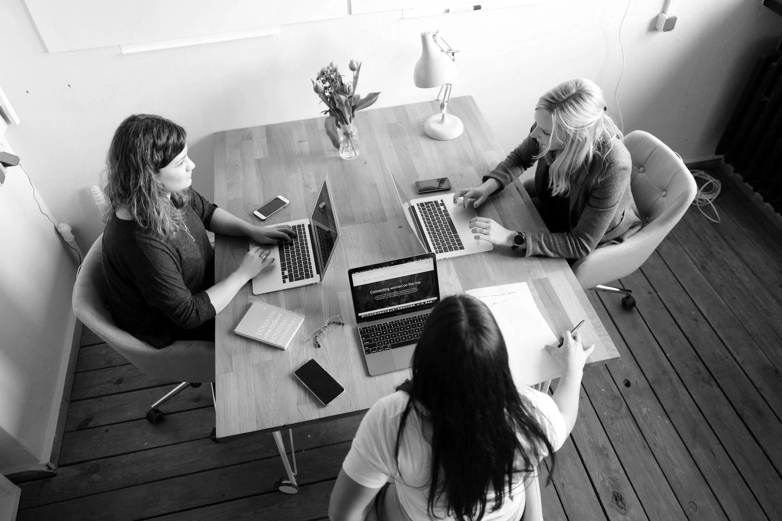 Groupe de femmes travaillant sur leurs ordinateurs portables dans une salle de réunion avec une table en bois et une décoration minimaliste, y compris une vase avec des fleurs et une lampe.