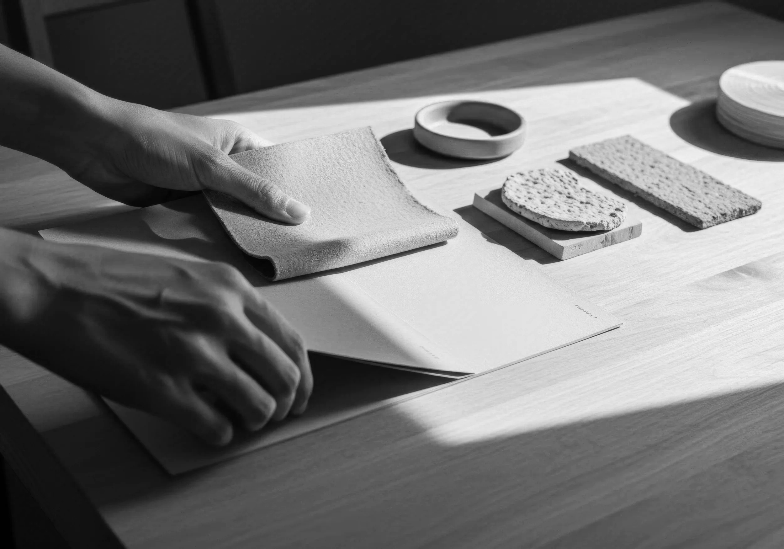 Main dans une main, tenant un morceau de tissu ou de matériau textile sur une surface en bois, avec des échantillons de matériaux variés, y compris des textures rugueuses et lisses, disposés sur la table.