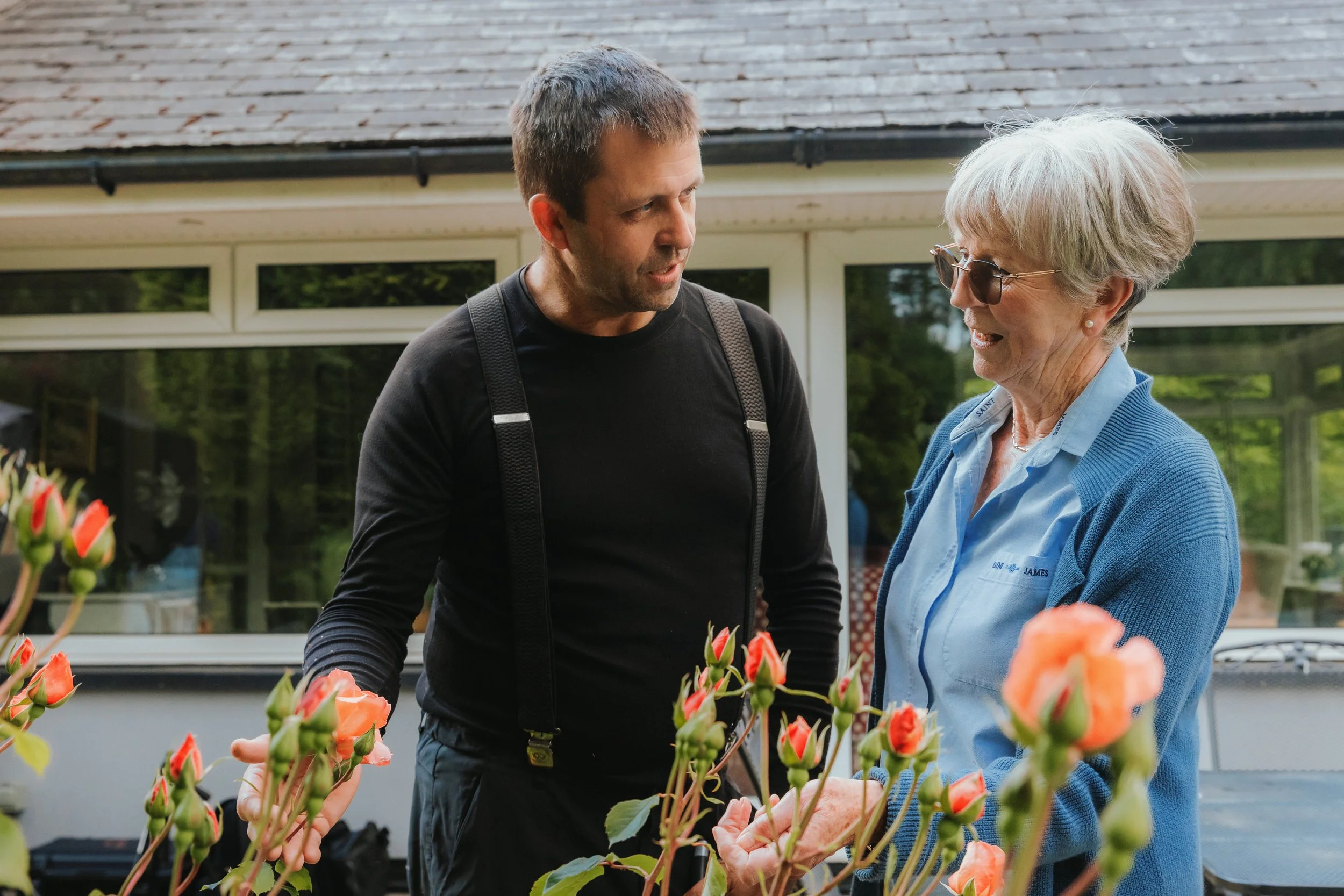 A carer and a woman smiling and chatting while tending to vibrant orange rose bushes in a garden.
