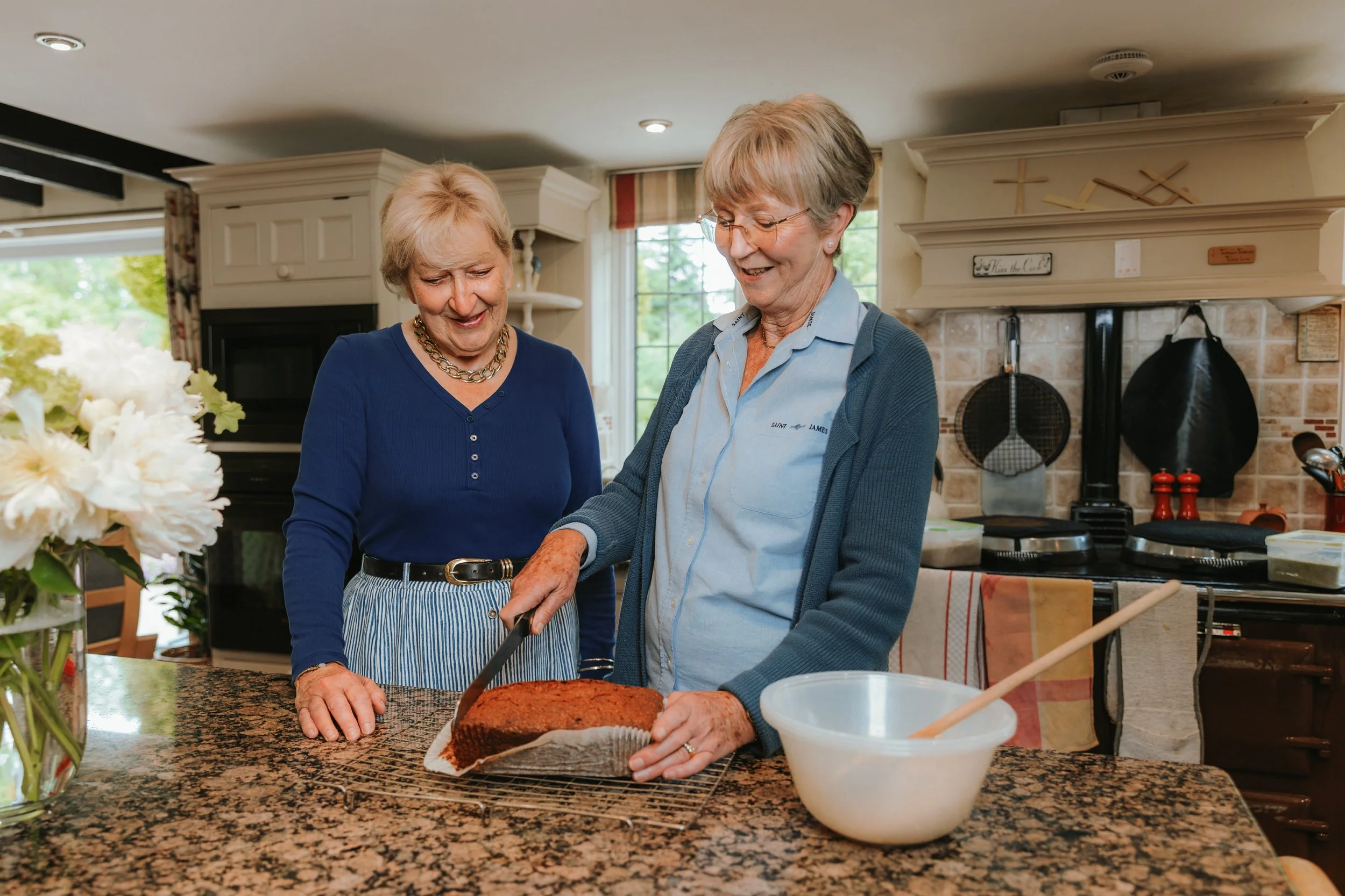 Two friends smiling and sharing a happy moment while slicing a loaf cake together in a kitchen.