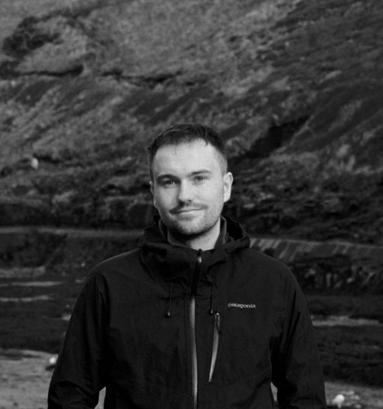 A young man with short dark hair and a slight smile, wearing a black Patagonia jacket, stands outdoors in front of a rugged mountain landscape in black and white.