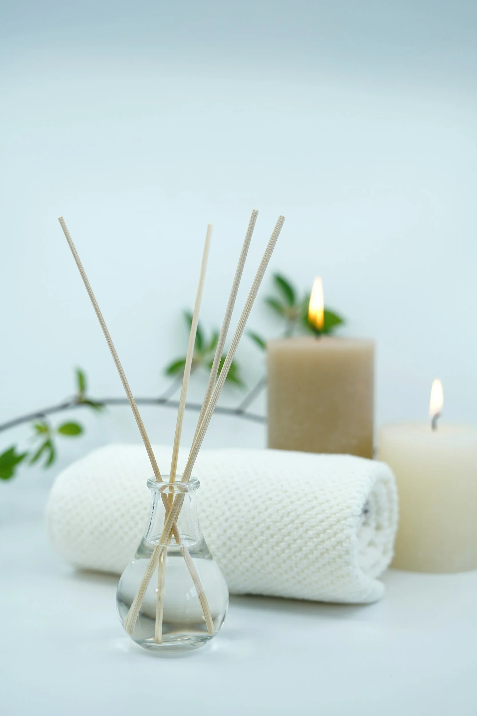 Empty glass vase with three wooden sticks, white textured cloth, blurred candles, plant leaves, and a soft blue background.