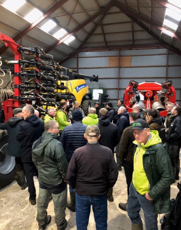Group of people inside a barn-like structure viewing large agricultural machinery, including a yellow tractor and a red planter.
