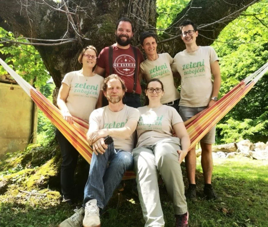 Six people, three women and three men, posing outdoors in front of a large tree with bright green leaves. They are standing and sitting near a colorful hammock, smiling at the camera. Some of the women are wearing T-shirts with the text 'Zeleni Zabojcek'.