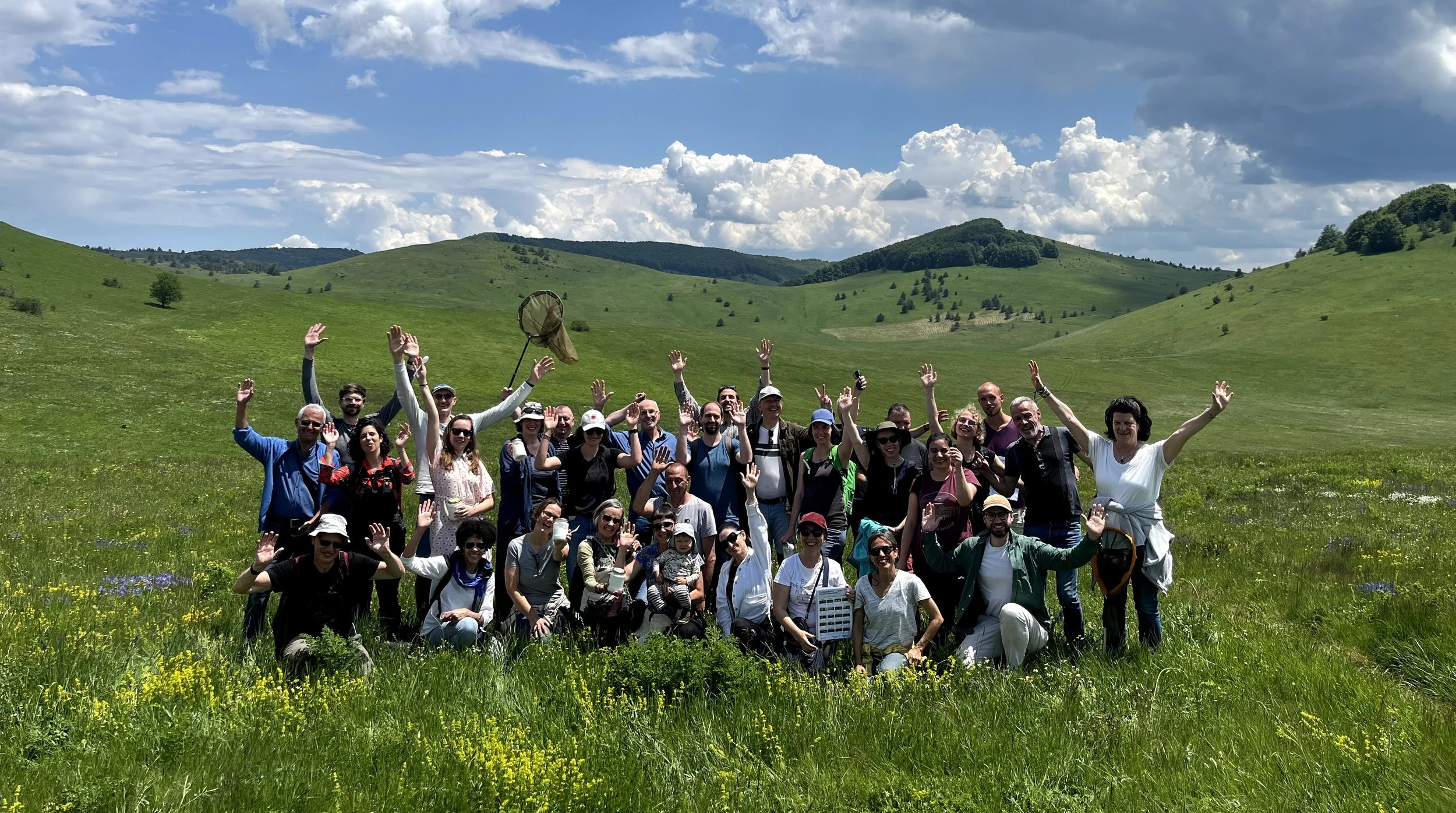 A large group of people posing together outdoors in a green grassy field with rolling hills and a partly cloudy sky in the background, many waving and smiling.