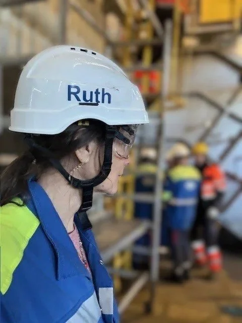 Close-up side view of a female construction worker wearing a white helmet labeled 'Ruth', safety glasses, and a colorful safety vest, with other workers and scaffolding in the background.