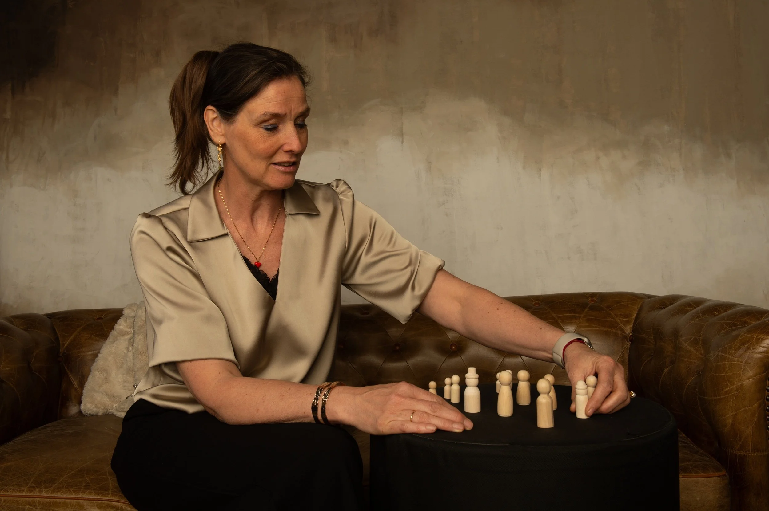 A woman with short brown hair, wearing a beige satin blouse, playing chess on a leather couch in a room with a plain textured wall.