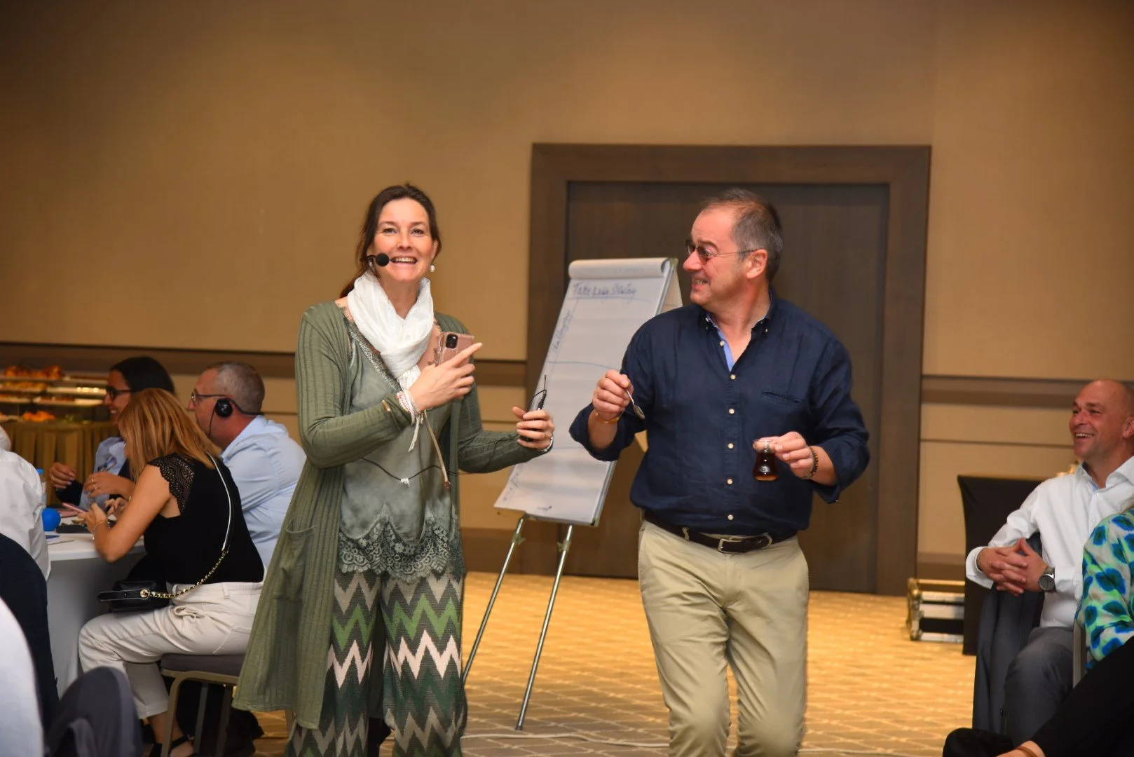 People smiling and talking in a conference or seminar room, some wearing headsets, with Ruth Verlinden holding a microphone and a man holding a glass of tea near a whiteboard.