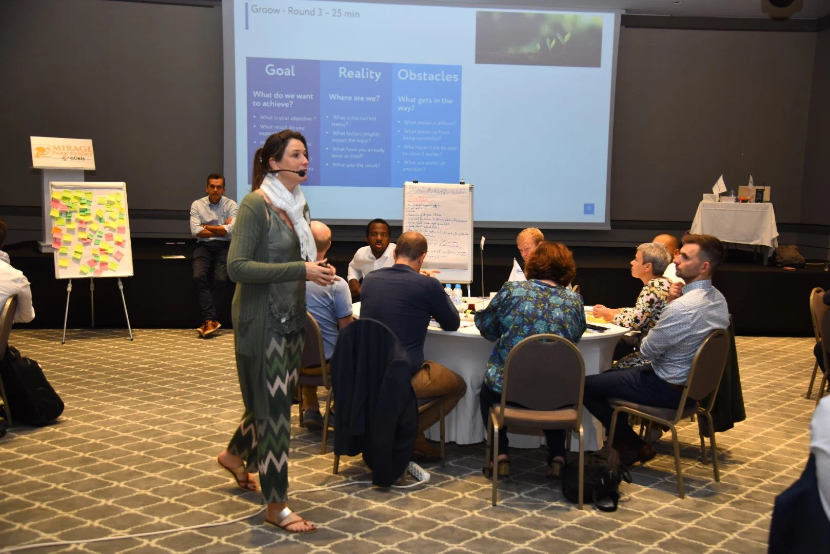 Ruth Verlinden is speaking to a group of people in a conference room. There is a large screen behind her displaying a presentation slide titled 'Goal, Reality, Obstacles'.