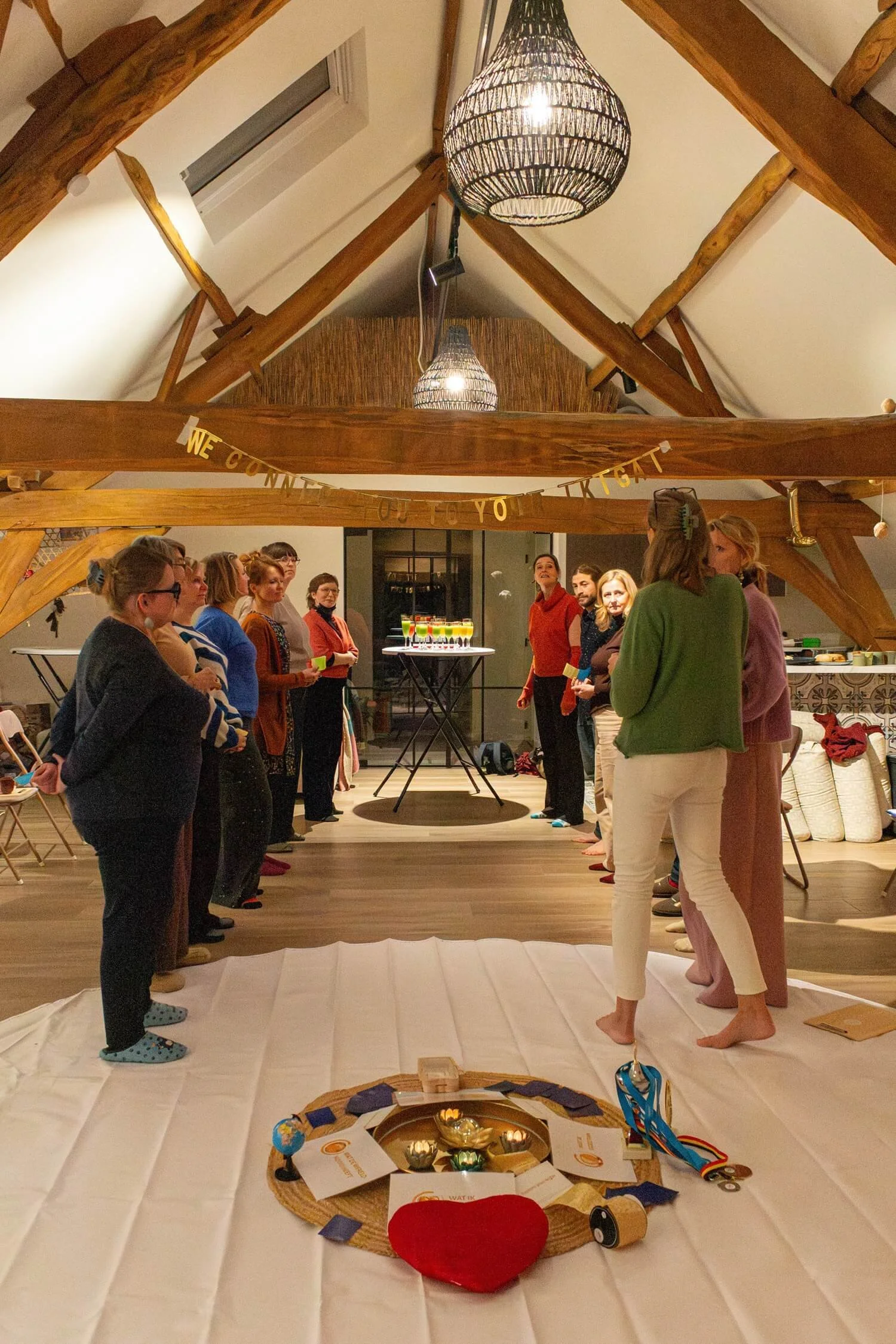A group of people standing in a circle indoors, with a decorated table and birthday party items in the foreground, celebrating a special occasion.