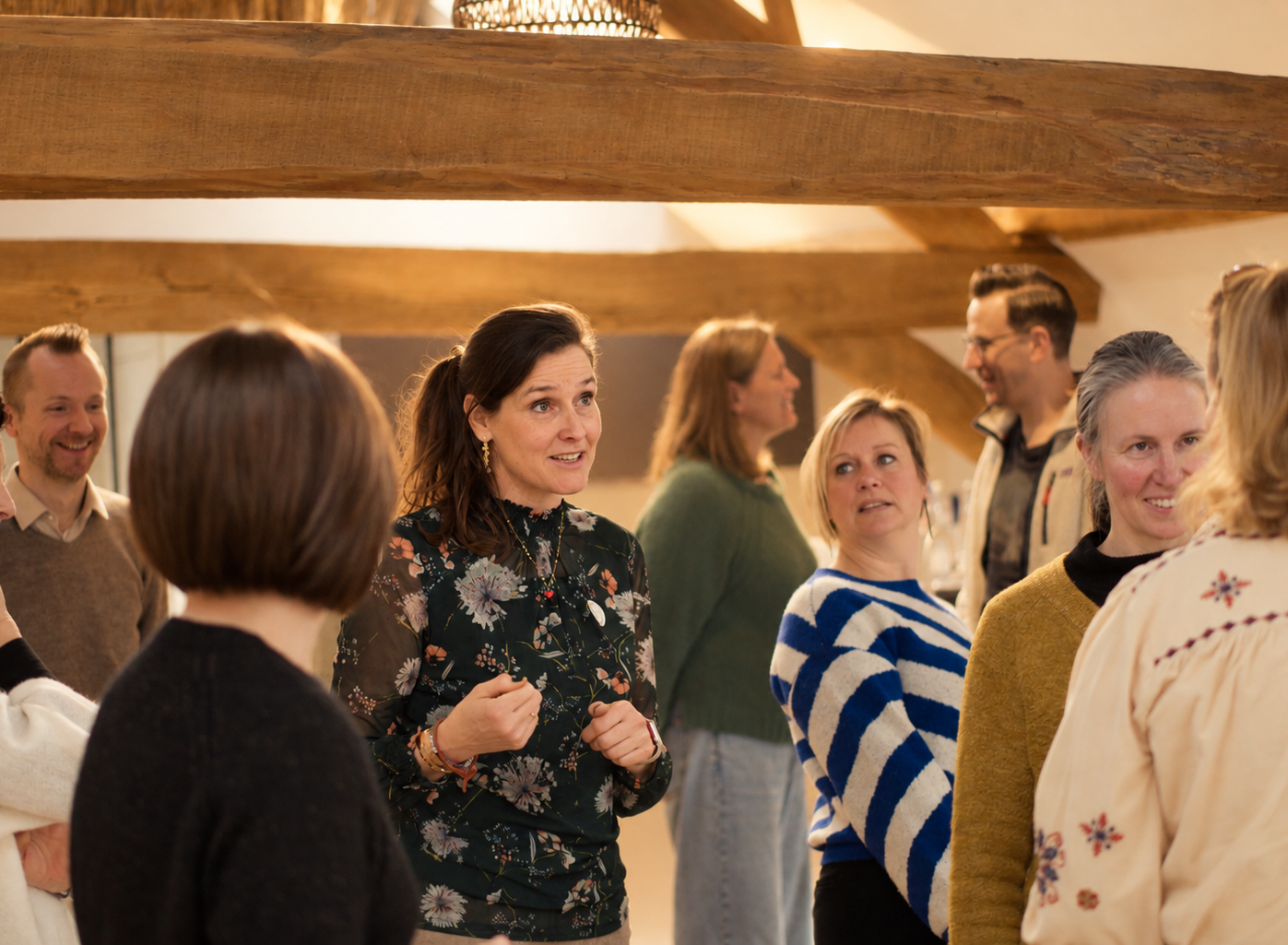 People gathered in a cozy, well-lit attic space with wooden beams, sitting on chairs and cushions, engaged in conversation and activities, with a large window at the back and a white cloth on the floor in the center.