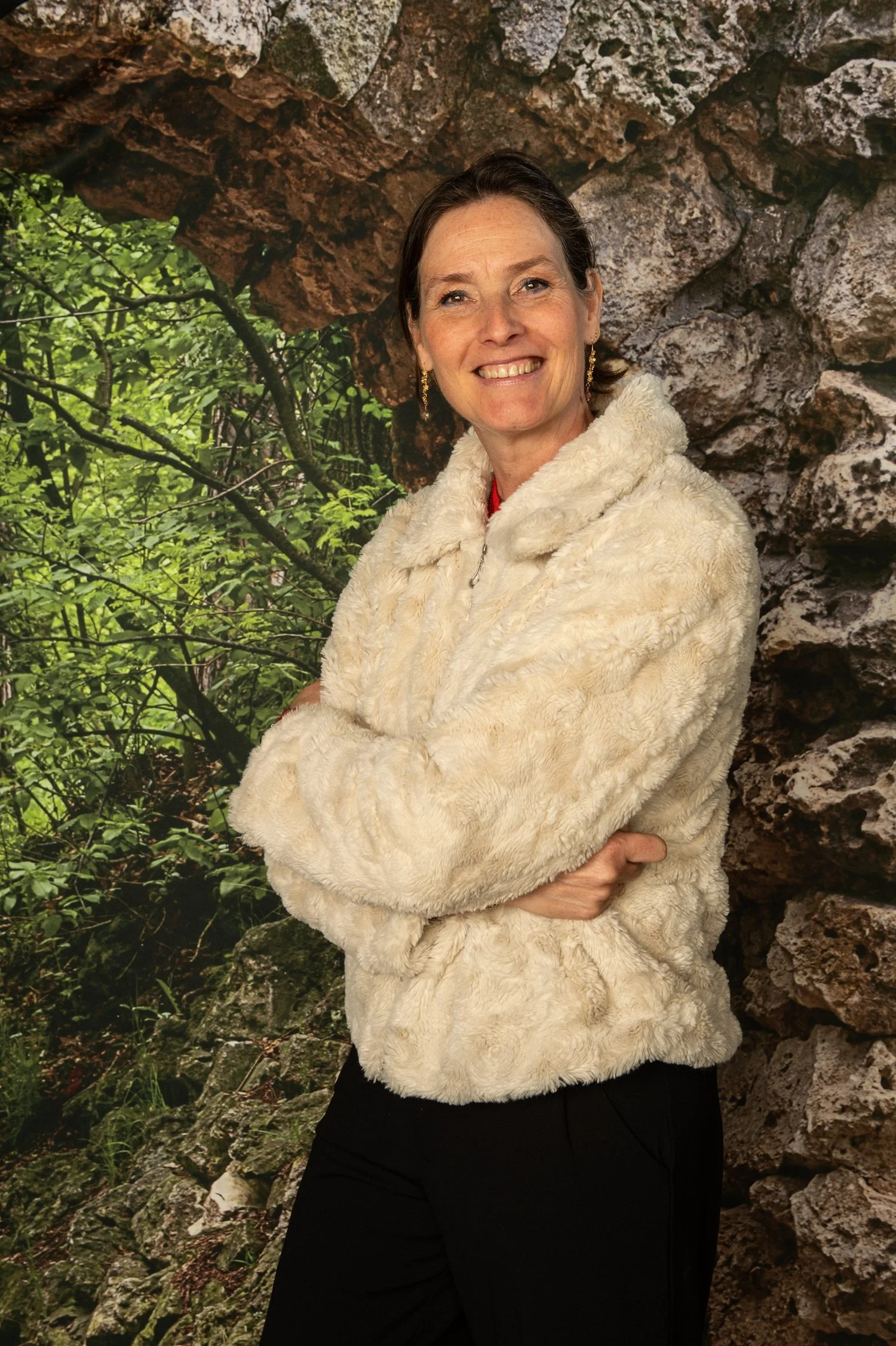A woman with brown hair, smiling, wearing a cream-colored fuzzy jacket, standing outdoors in front of a rocky and green forest background.