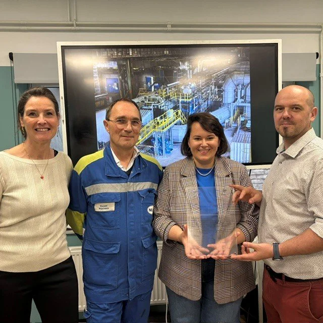 Four people standing in front of a large screen displaying industrial equipment indoors, with two women and two men smiling. The woman on the right is holding a clear glass award.