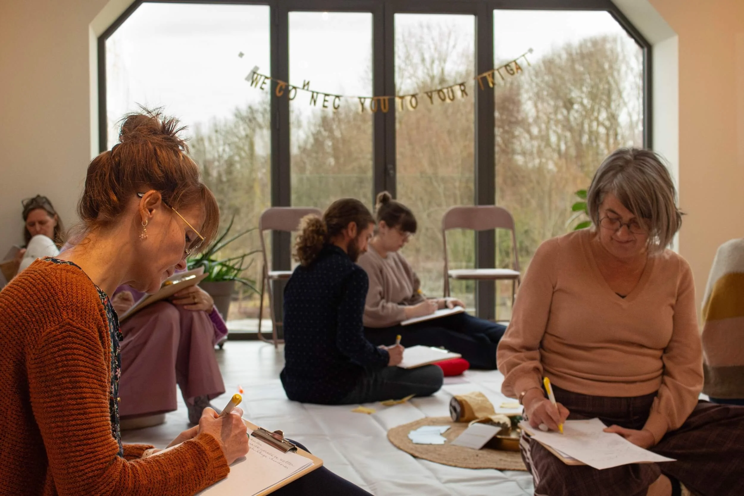 Group of women sitting on the floor with notebooks, pens, and papers, participating in a workshop or class, with a large window in the background.