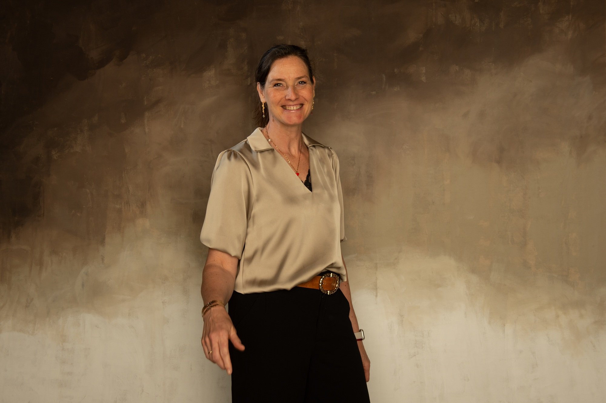 A woman with short dark hair, wearing a beige satin blouse, black pants with a decorative belt, standing against a textured, painted wall. She is smiling at the camera, accessorized with earrings, a necklace, bracelets, and a wristwatch.