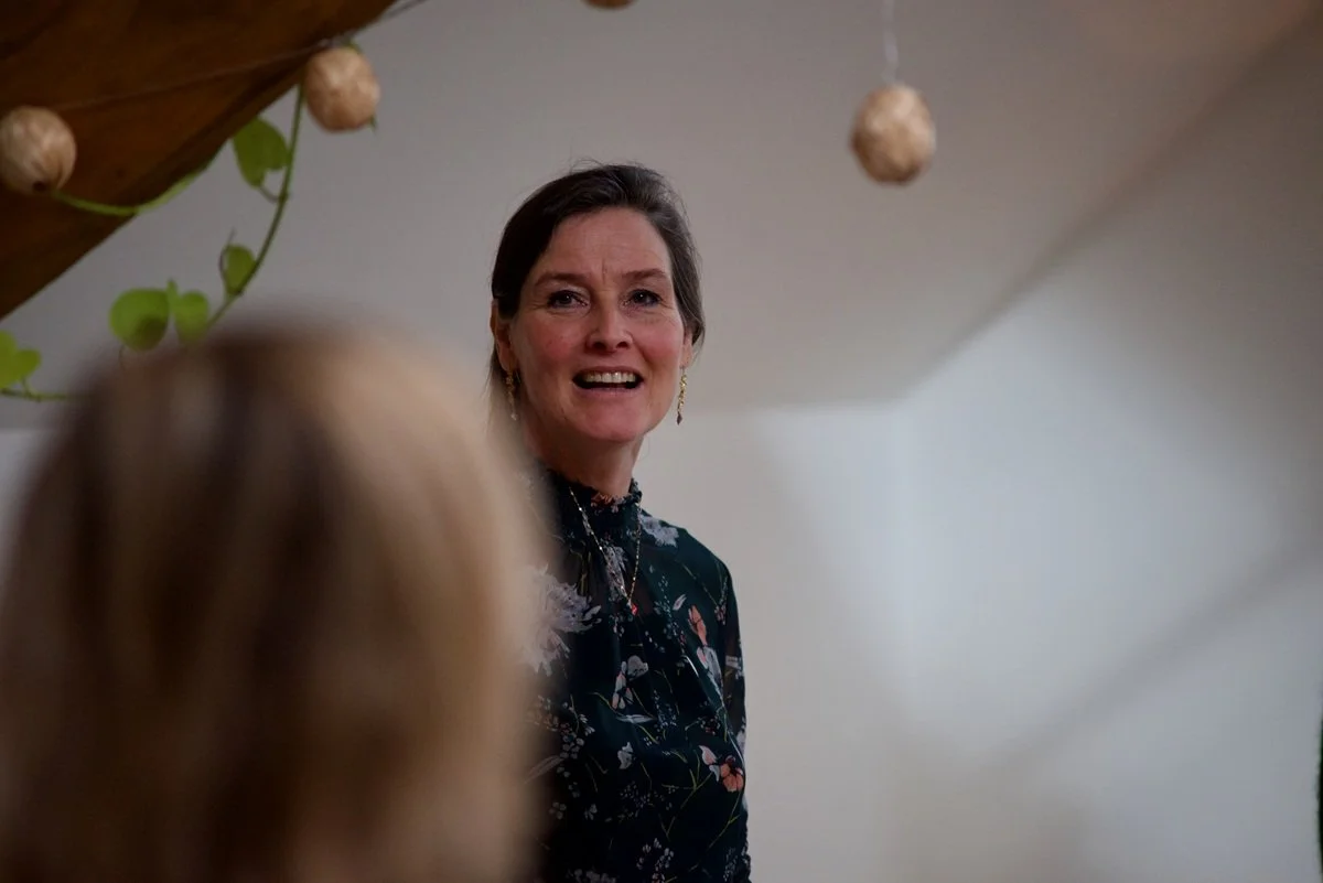 A woman with short dark hair, wearing a dark floral dress, smiling and talking to a person with blonde hair, in a cozy home setting decorated with hanging paper lanterns and green plants.
