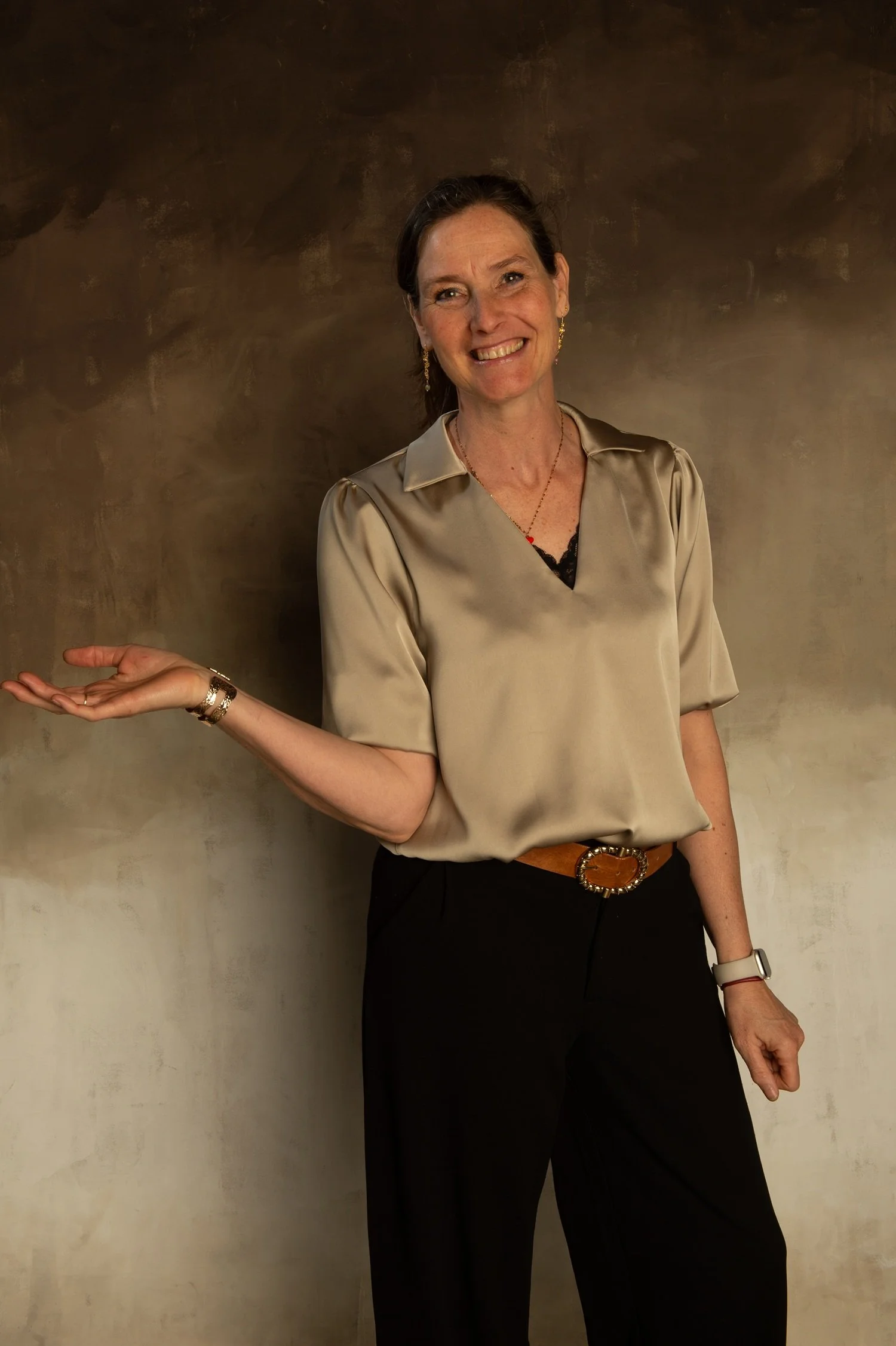 Ruth Verlinden, transformation coach,  standing against a dark background, wearing a beige silky blouse, black pants with a gold belt, and jewelry, smiling, with one hand extended outward.
