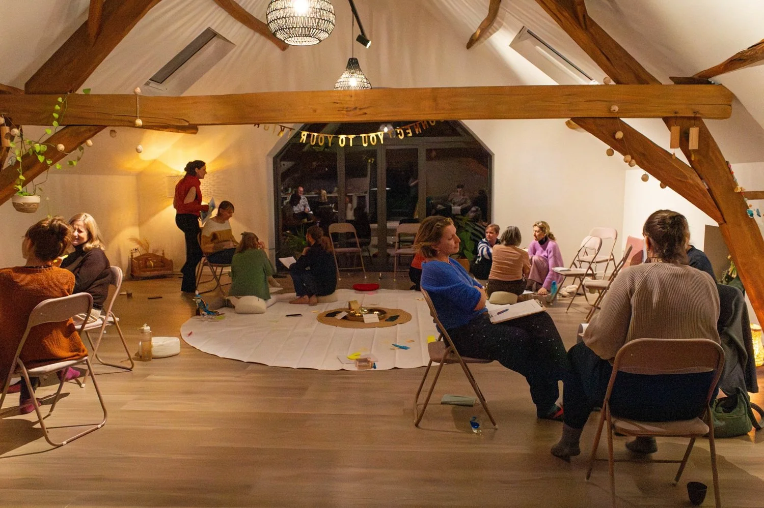People gathered in a cozy, well-lit attic space with wooden beams, sitting on chairs and cushions, engaged in conversation and activities, with a large window at the back and a white cloth on the floor in the center.
