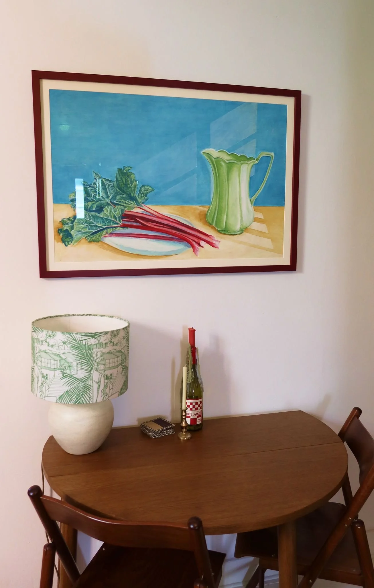 A wooden dining table with four matching chairs, a decorative lamp with a green leafy pattern, a candle holder, a bottle, and a small jewelry box. Above, a colorful painting of a green pitcher and rhubarb on a table is hung on the wall.