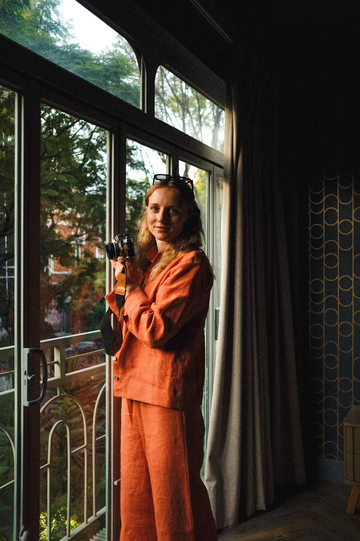 A woman in an orange suit holding a camera stands near a large window with trees outside.