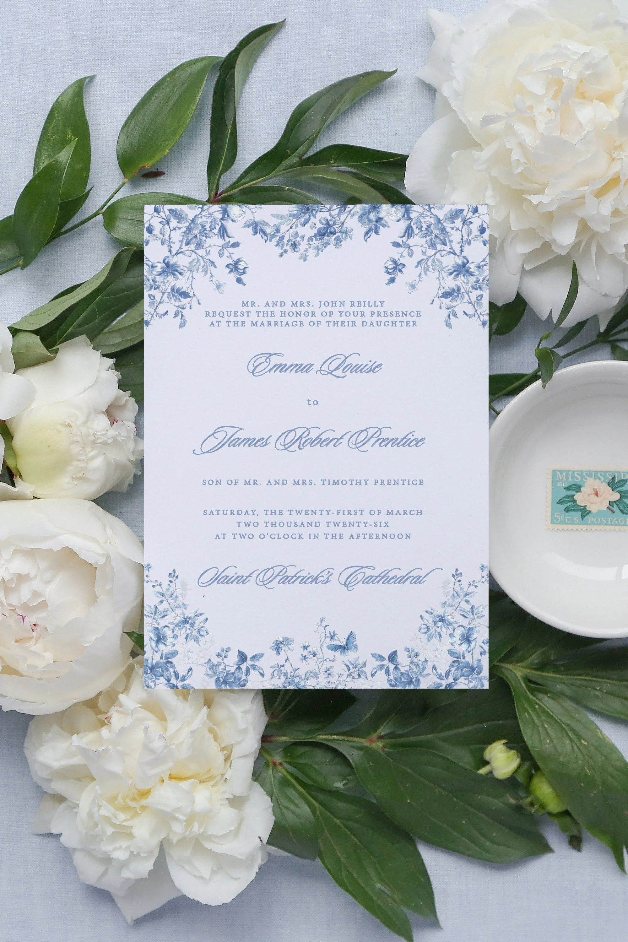beautifully designed wedding inviation including chinoiserie blue floral designs and script text. Laying on top of white peonies.