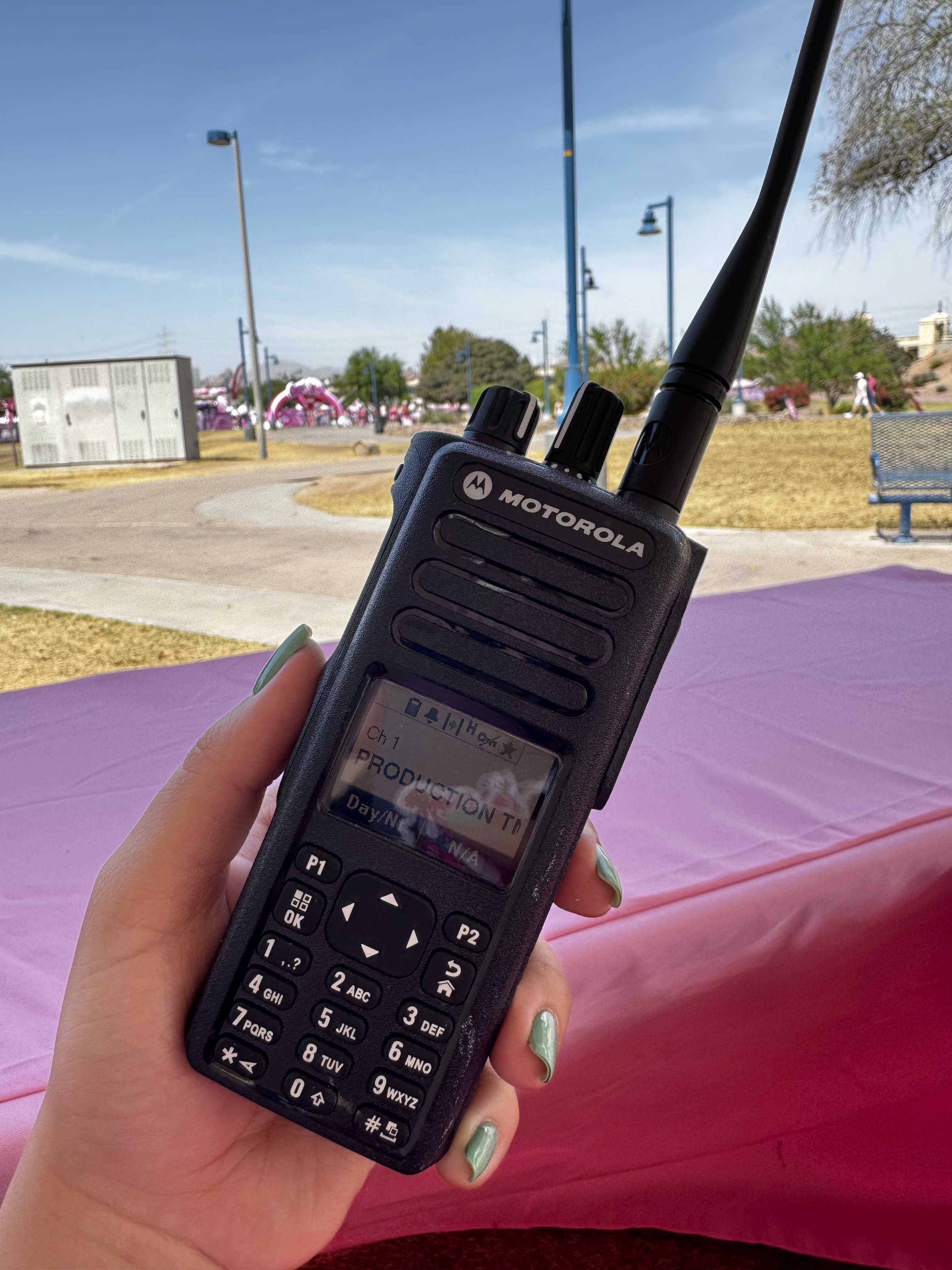 Close-up of a Motorola walkie-talkie held by a person's hand, with a scenic park background featuring pathways, benches, trees, and a pink inflatable structure in the distance, under a clear blue sky.
