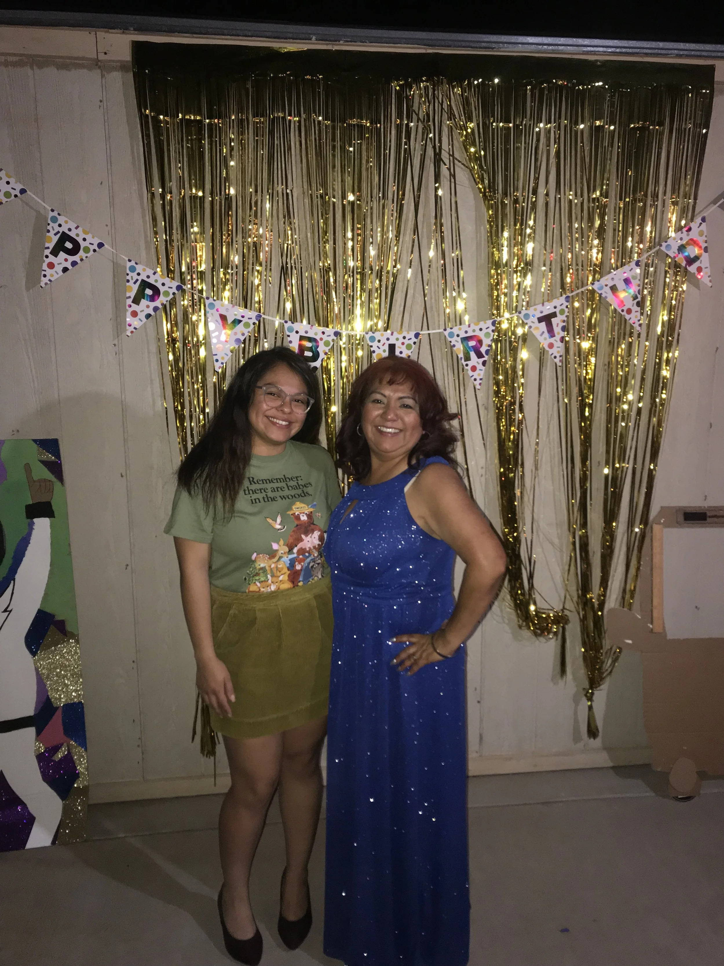 A woman in a blue sequined dress and a girl in a green t-shirt and yellow skirt pose together in front of a gold tinsel backdrop at a celebration, with colorful 'Happy Birthday' banners hanging above them.