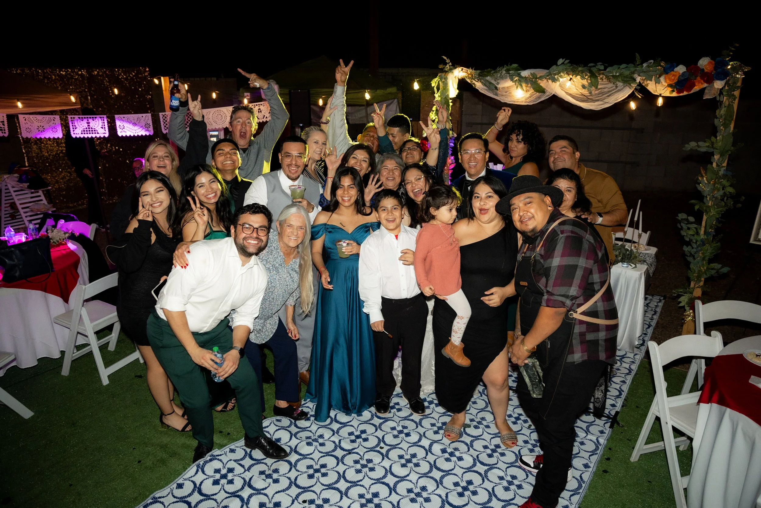 A large group of people celebrating at an outdoor party at night. They are smiling, waving, and posing for the camera under decorative lights and floral decorations. Some hold drinks, and there are tables with chairs and tablecloths nearby.