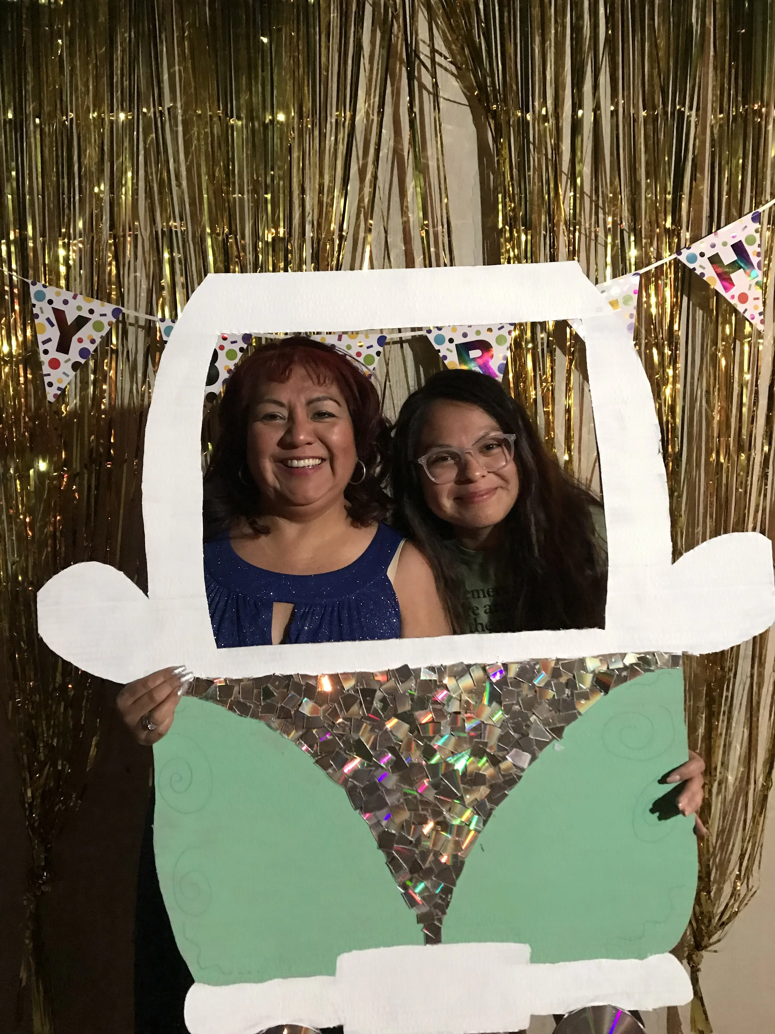 Two women holding a large cutout of the Superman logo. One woman has short red hair and is wearing a blue dress, the other has long dark hair, glasses, and is wearing a green shirt. They are in front of a gold tinsel backdrop with a colorful banner reading 'YAY' and confetti decorations.