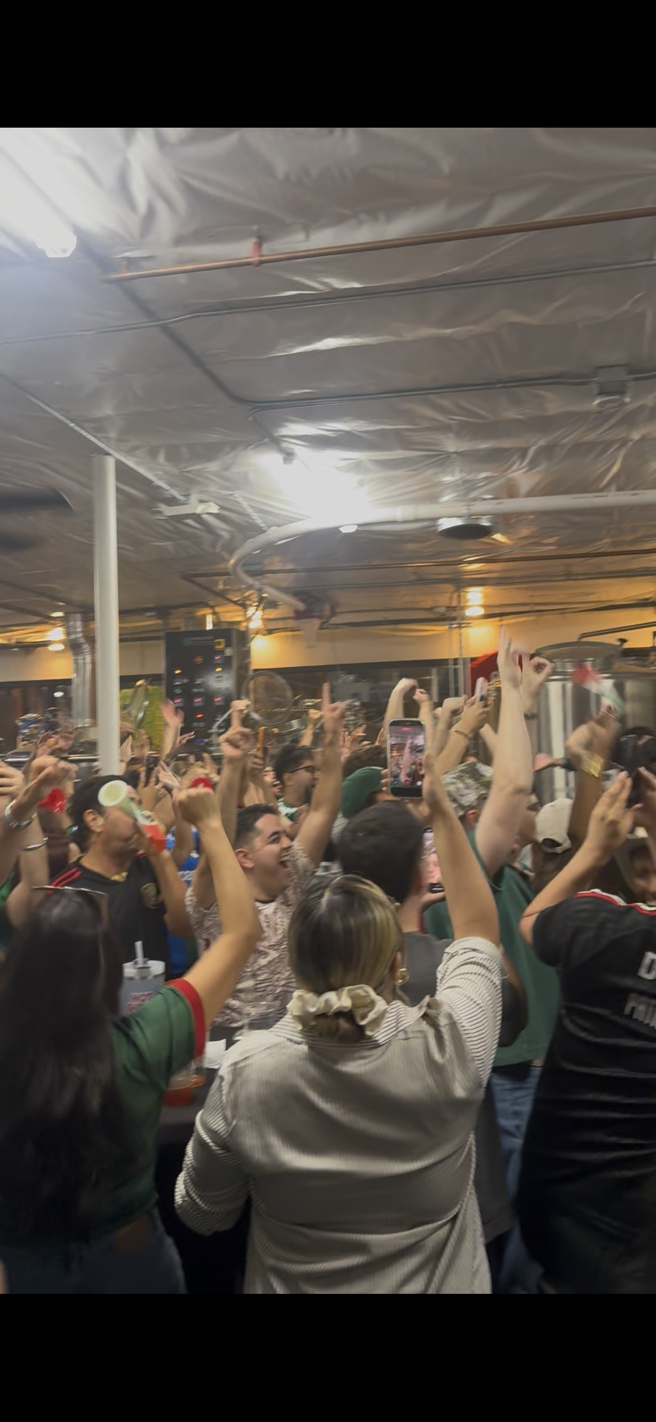 A large crowd of people enjoying a lively event in an indoor setting, some holding smartphones and raising their hands.