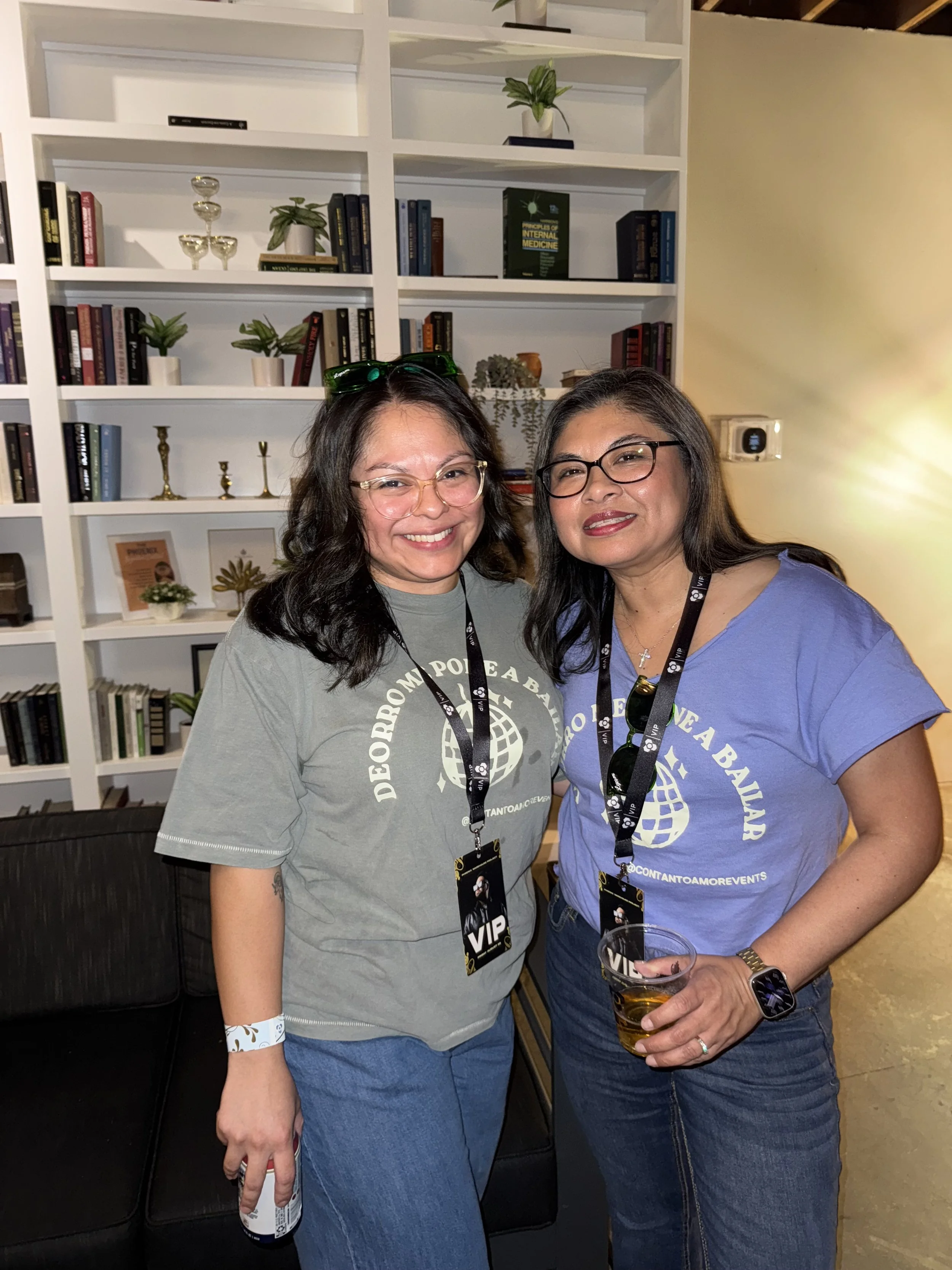Two smiling women with glasses, wearing gray and blue t-shirts, standing in front of a white bookshelf filled with books, plants, and decorative items, at a social event.