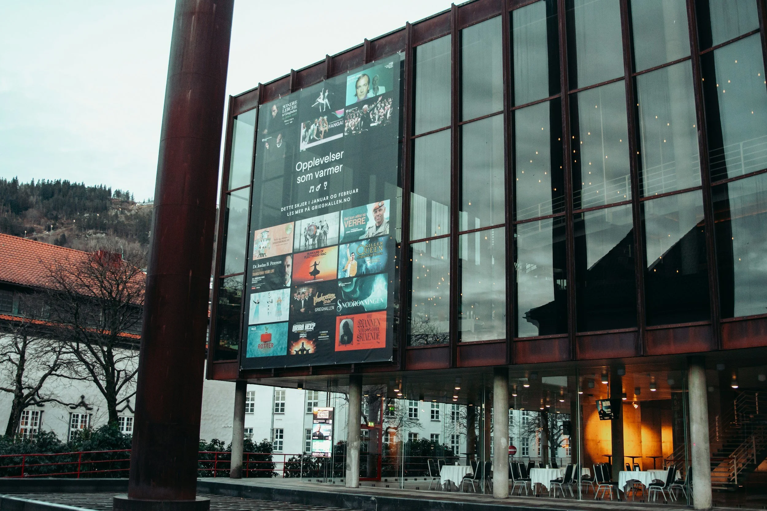 A modern building with large glass windows displaying posters of musicals and performances, outdoor seating with tables and chairs, trees, and a cloudy sky.
