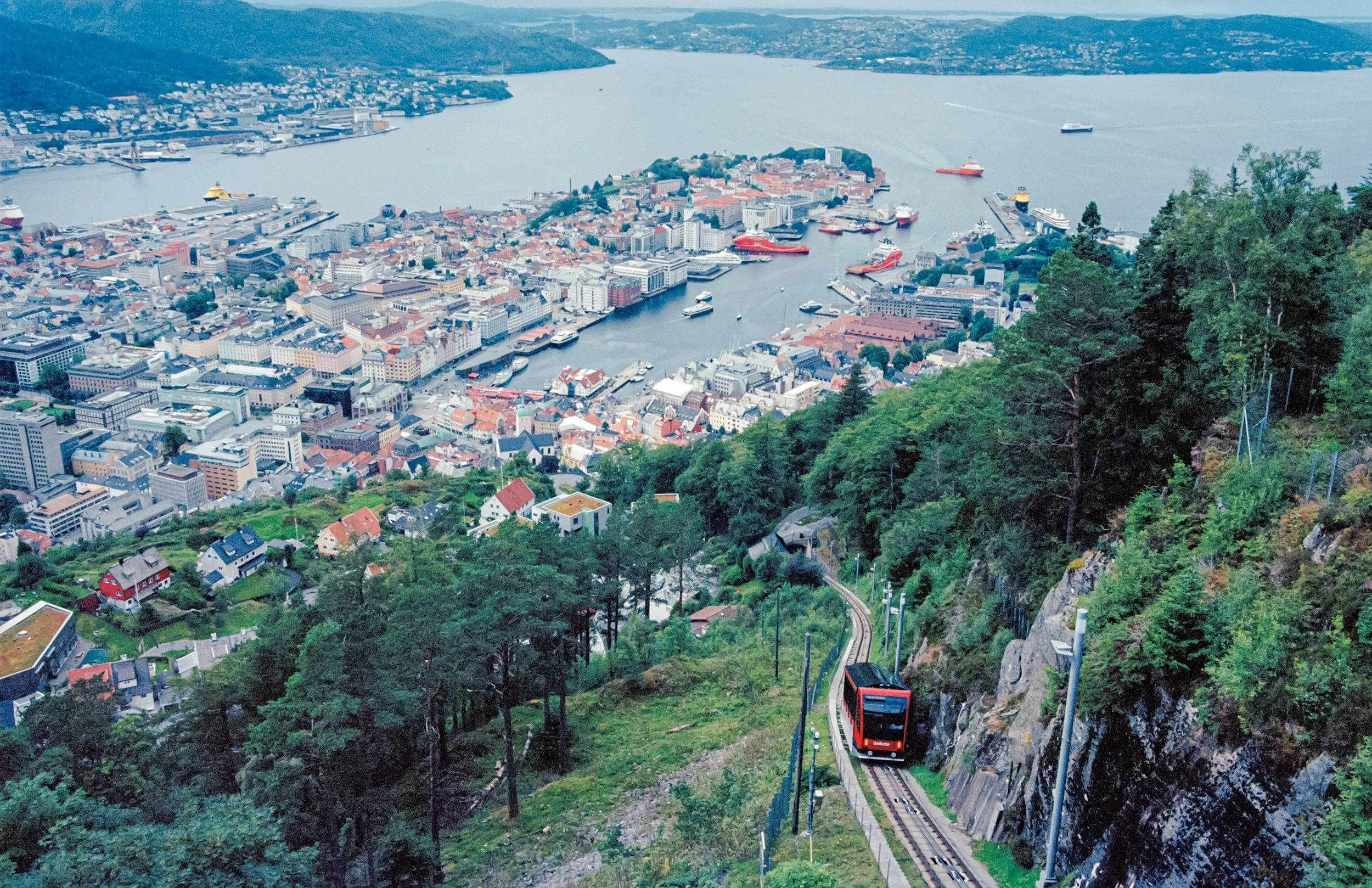 Aerial view of a city by a large lake with docks and boats, surrounded by green hills and forests, with a funicular railway ascending a steep hillside in the foreground.