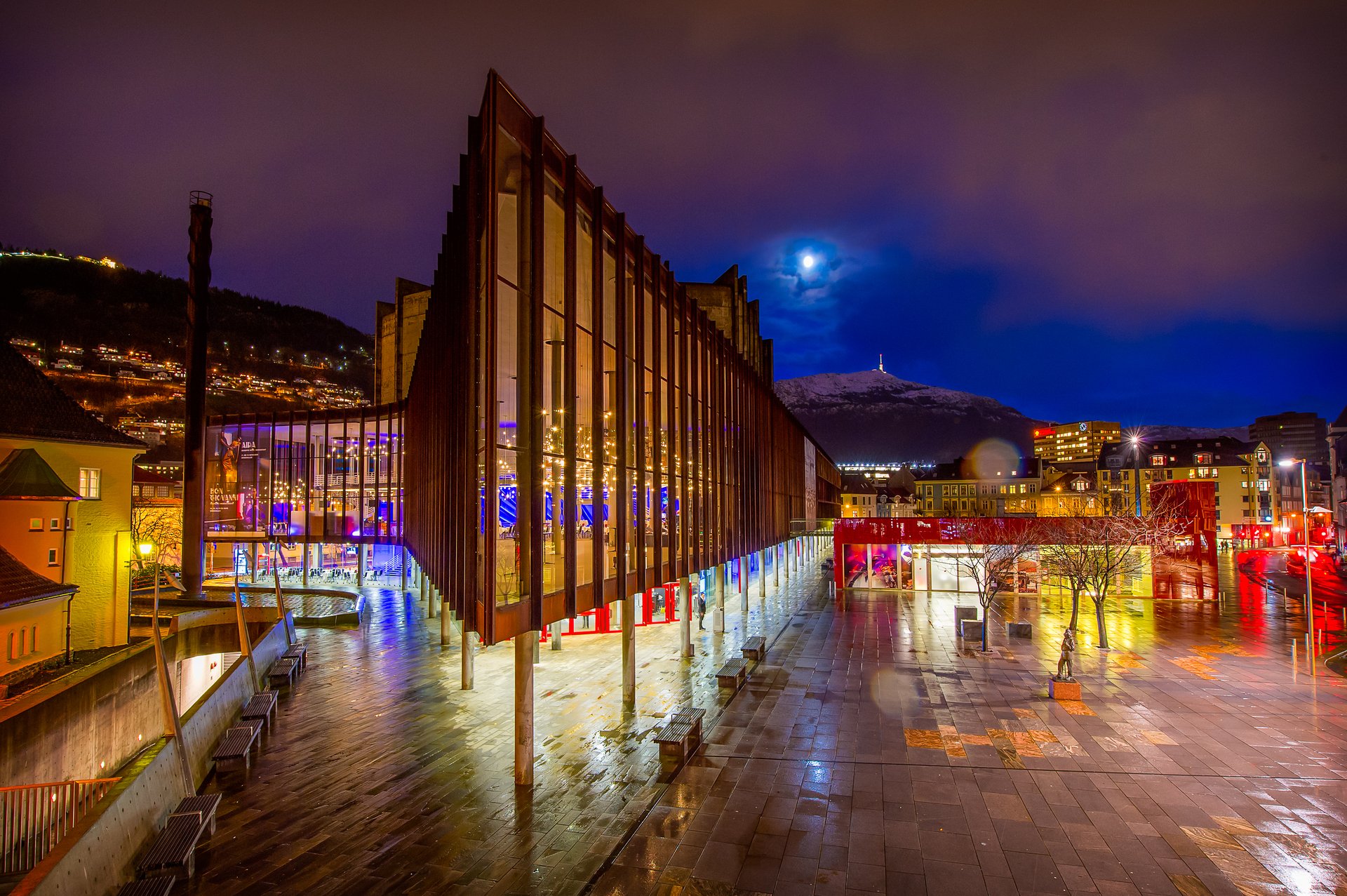 Nighttime cityscape with a modern glass building illuminated from within, wet pavement reflecting lights, hills with houses in the background, a mountain with a tower on top, and a moon partially obscured by clouds.