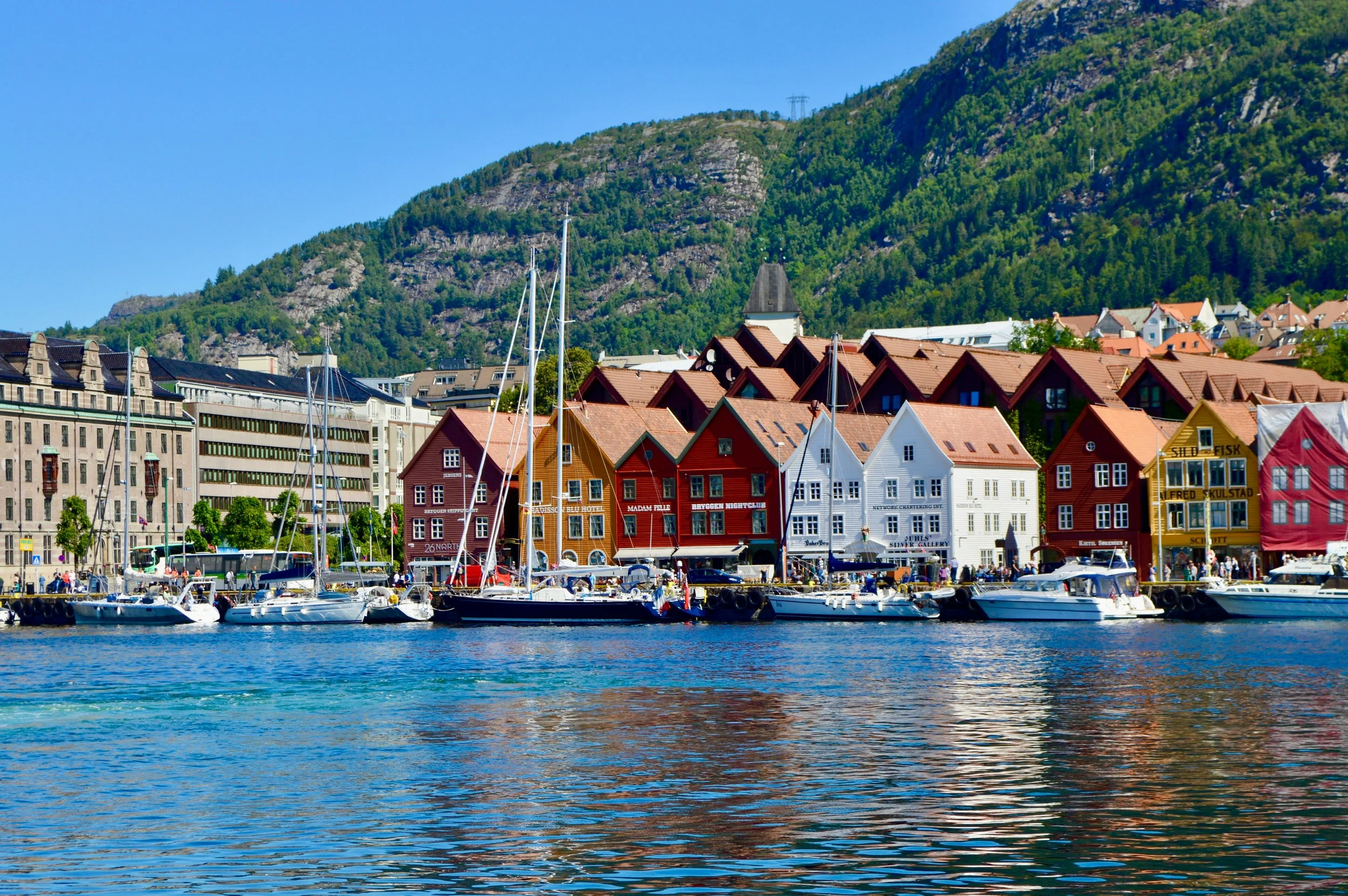 A harbor with boats docked, colorful buildings along the waterfront, lush green hills in the background, and a clear blue sky.