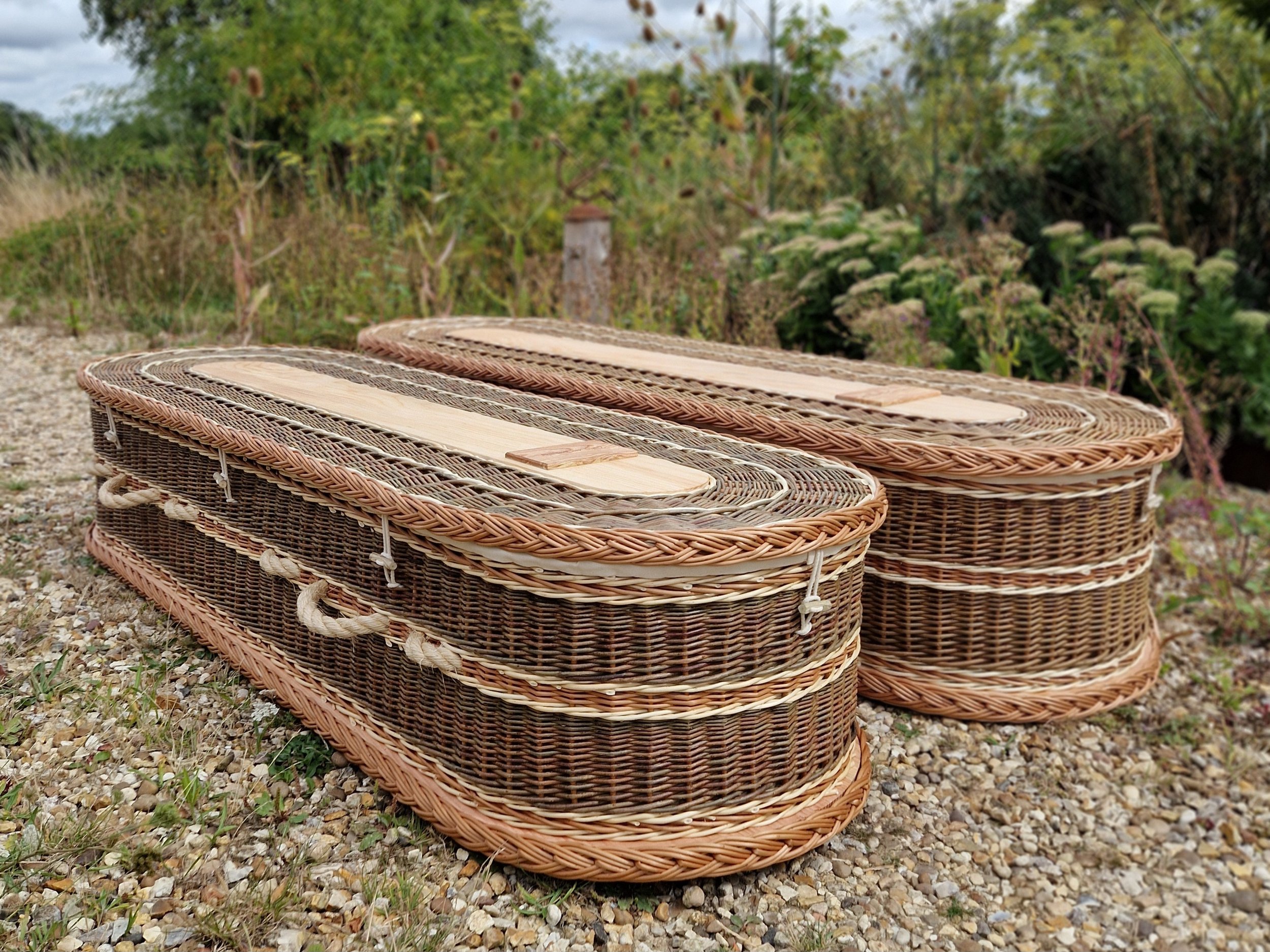 Stacked wicker coffins with wooden lids outdoors on gravel ground, greenery and trees in background.
